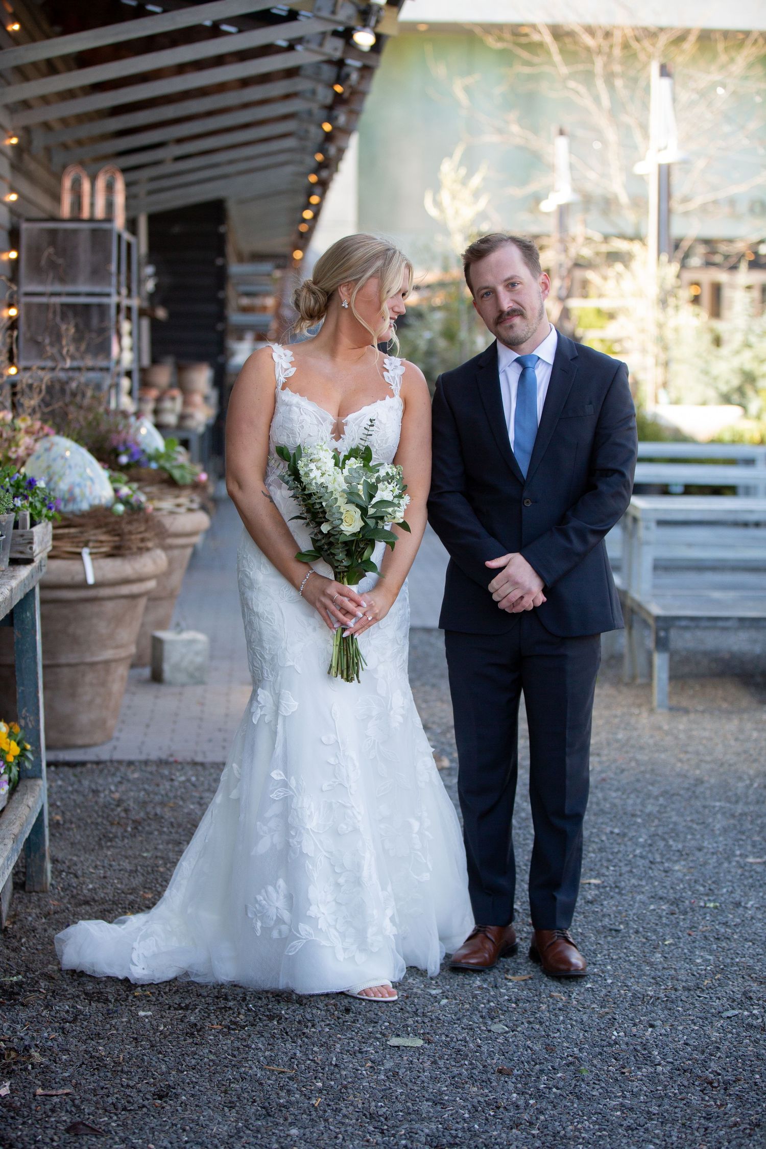 Bride in lace mermaid gown holding white bouquet stands with groom in navy suit and blue tie at outdoor wedding venue.
