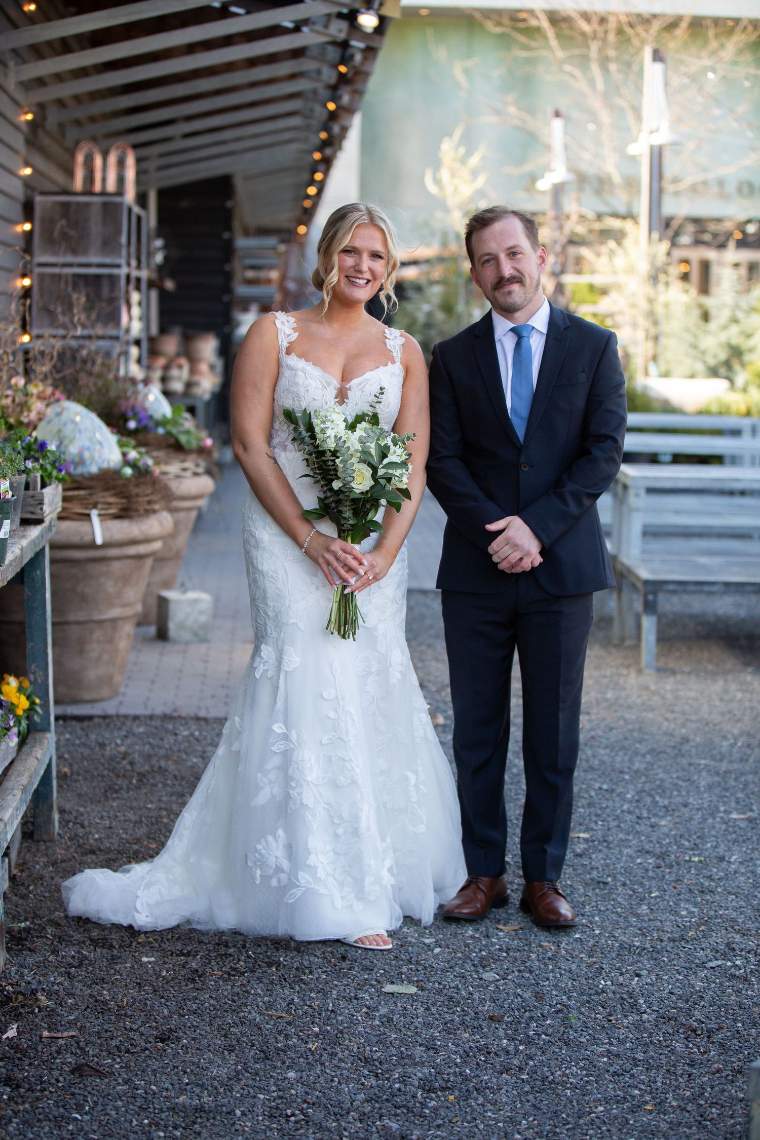 Bride in white lace gown holding bouquet stands with groom in navy suit at outdoor wedding venue.