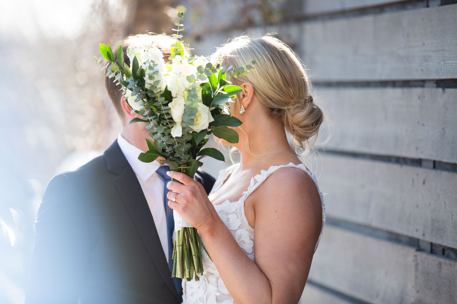 Bride and groom sharing a romantic kiss hidden behind a white floral wedding bouquet outdoors.