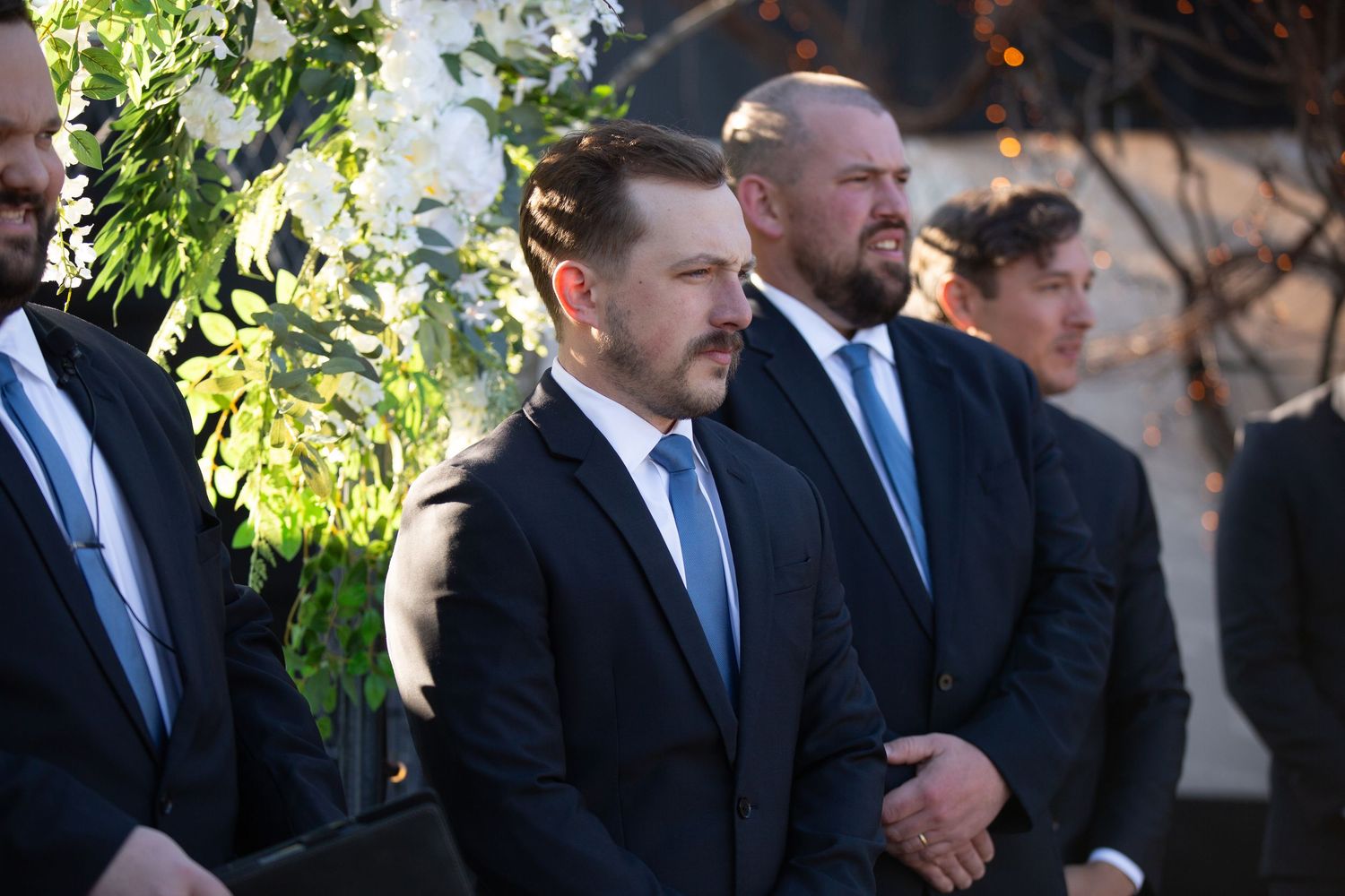 Men in dark suits standing outdoors at a formal event, possibly a wedding ceremony with white floral arrangements.