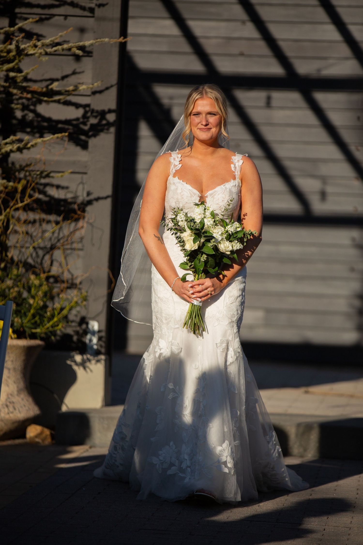 Bride in lace mermaid wedding dress holding white floral bouquet with veil, standing outdoors in sunlight.