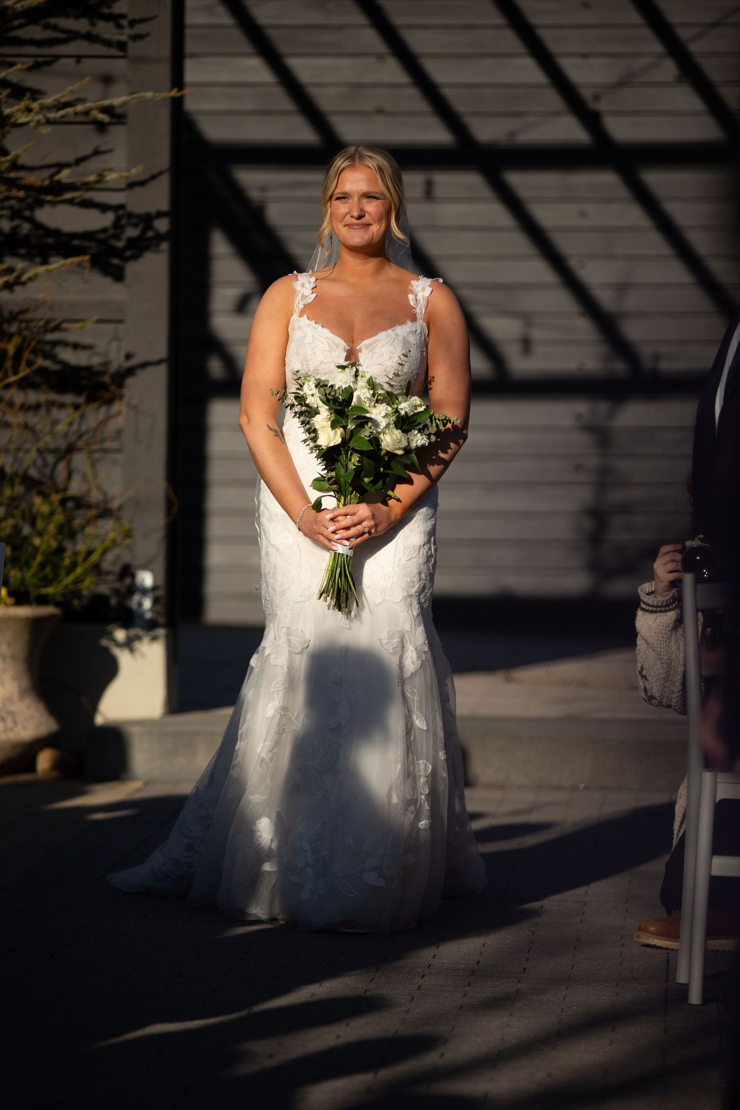 Bride in white lace mermaid gown holding white floral bouquet standing outdoors in sunlight at wedding ceremony.