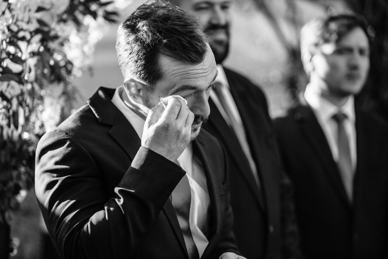 Emotional groom wiping tears at wedding ceremony in black and white photo with groomsmen in background.