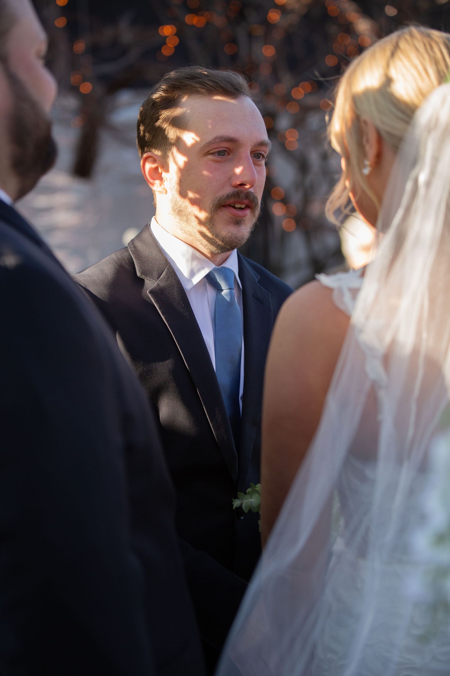 Groom in dark suit and blue tie exchanges vows with blonde bride in white veil at outdoor wedding ceremony.