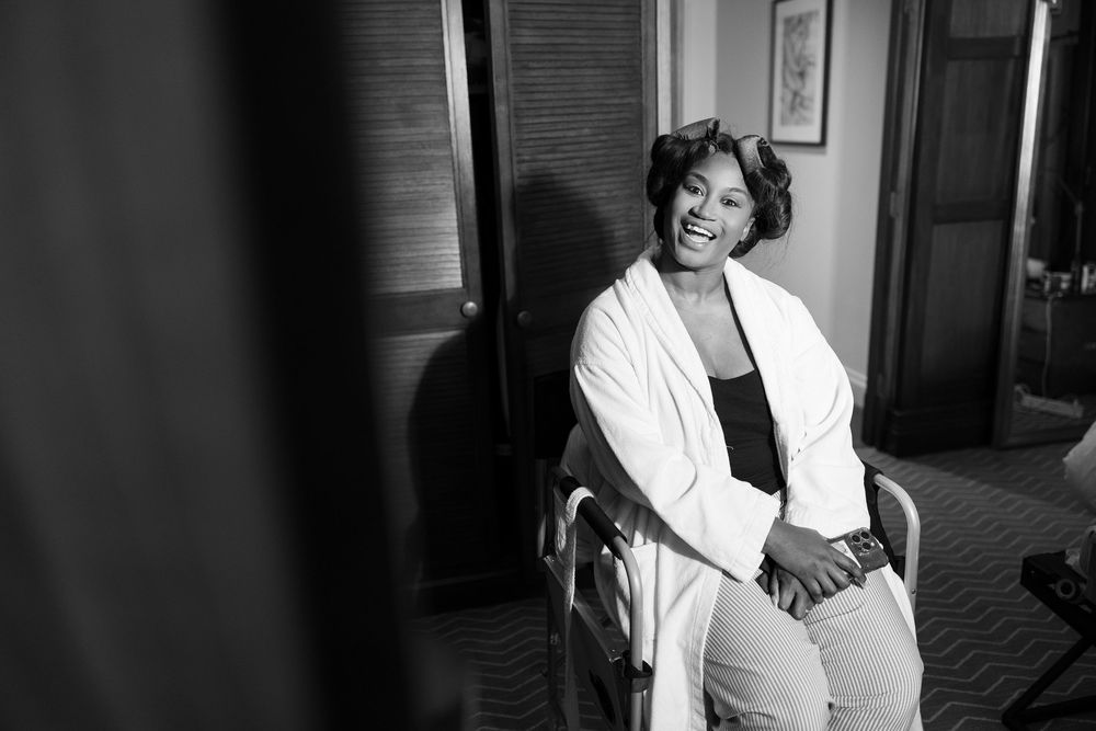 Black and white photo of smiling Black woman in white robe sitting in chair during wedding preparations.