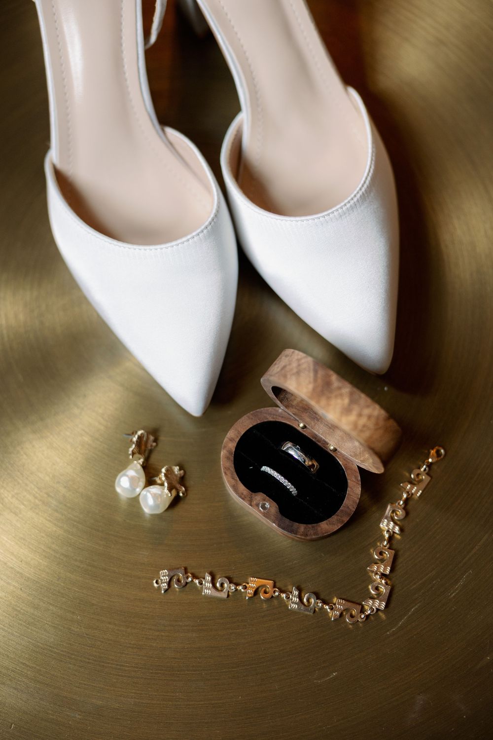 White bridal heels with pearl earrings, gold bracelet, and wooden ring box on a gold surface.