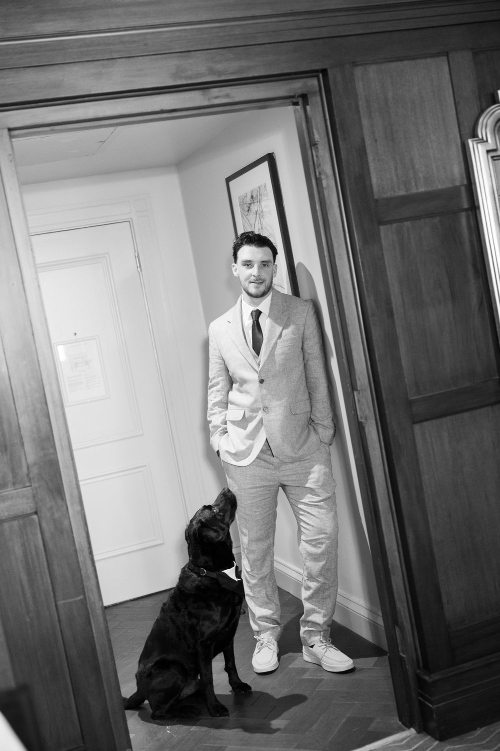 Black and white portrait of a groom in a suit standing in a doorway with a black Labrador dog.