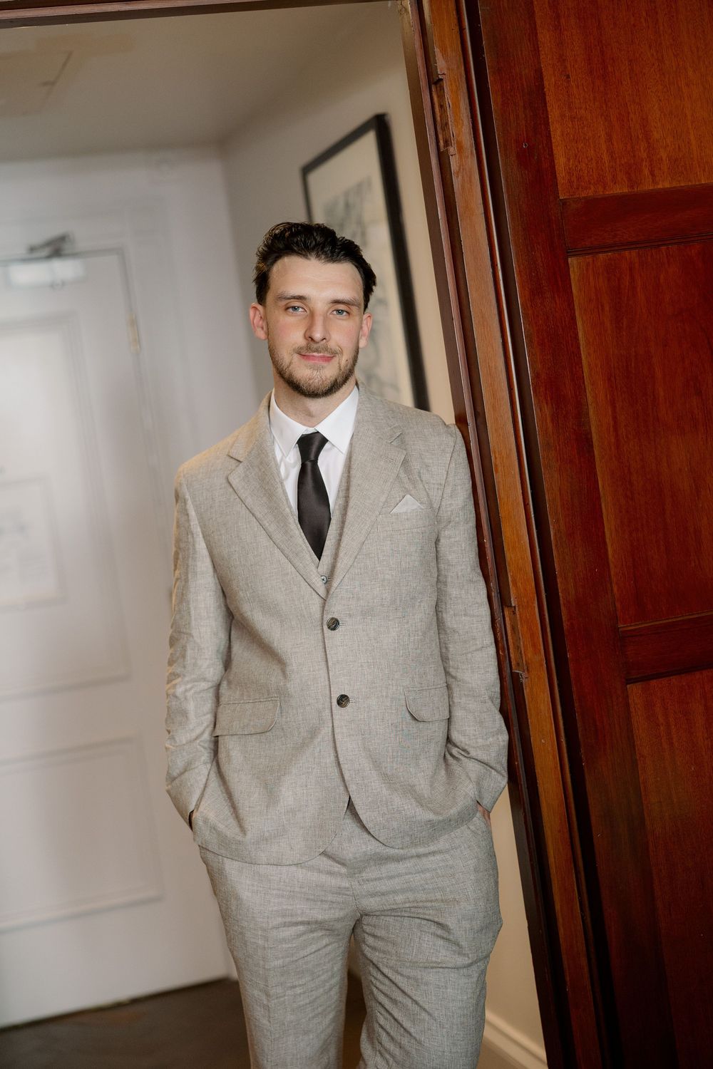 A groom in a light gray three-piece suit with black tie stands confidently by a wooden door.