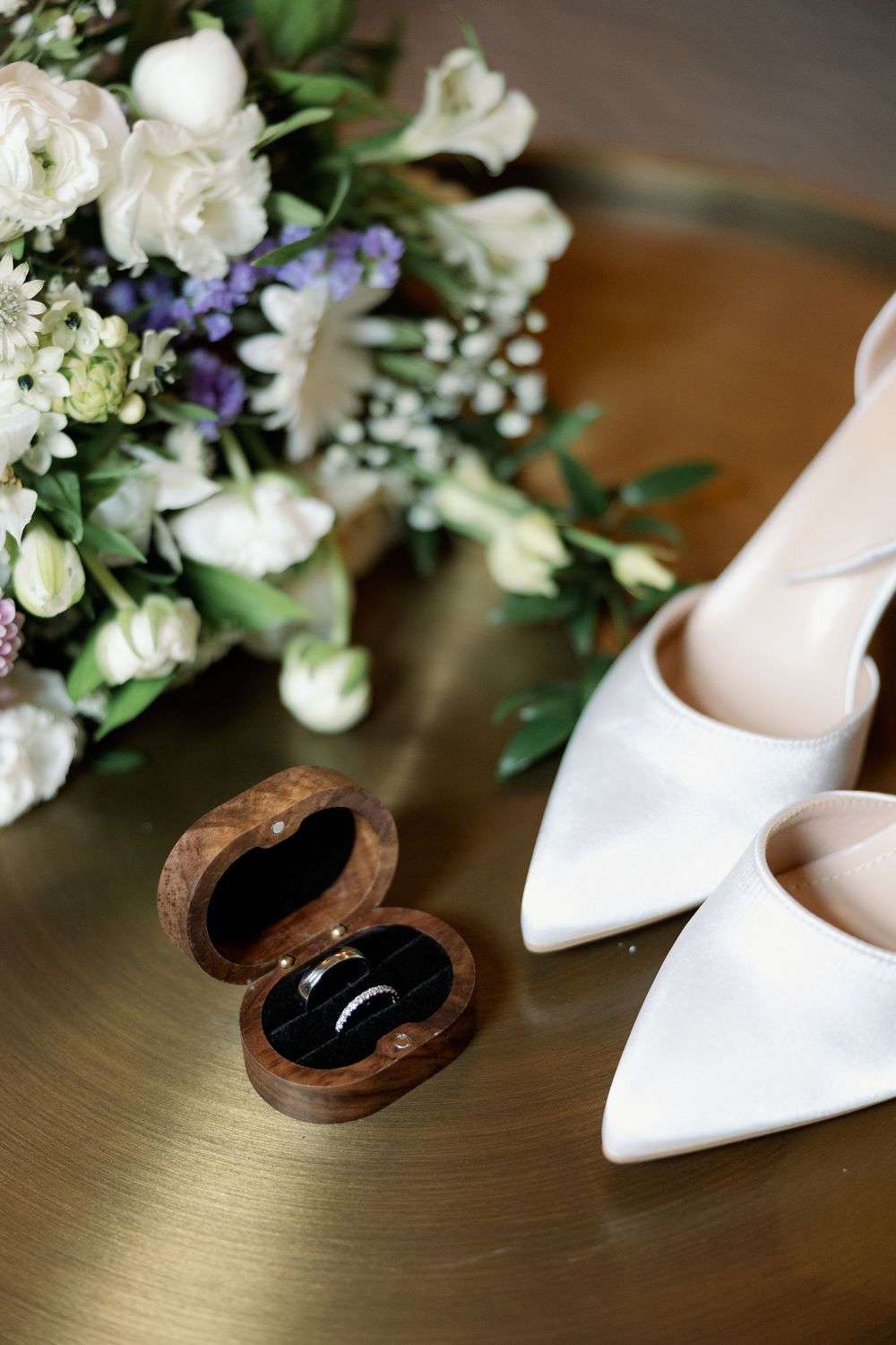Wedding flat lay featuring white bridal heels, wooden ring box with bands, and a white floral bouquet.