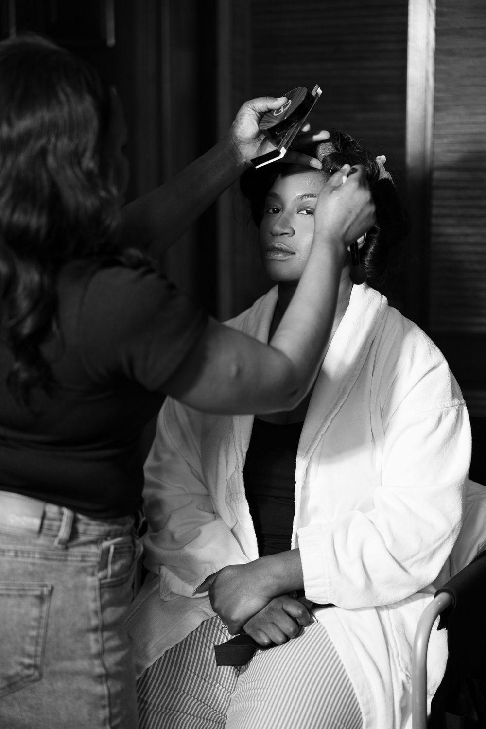 Black and white photo of a bride having her hair styled by a hairdresser while seated in a white robe.