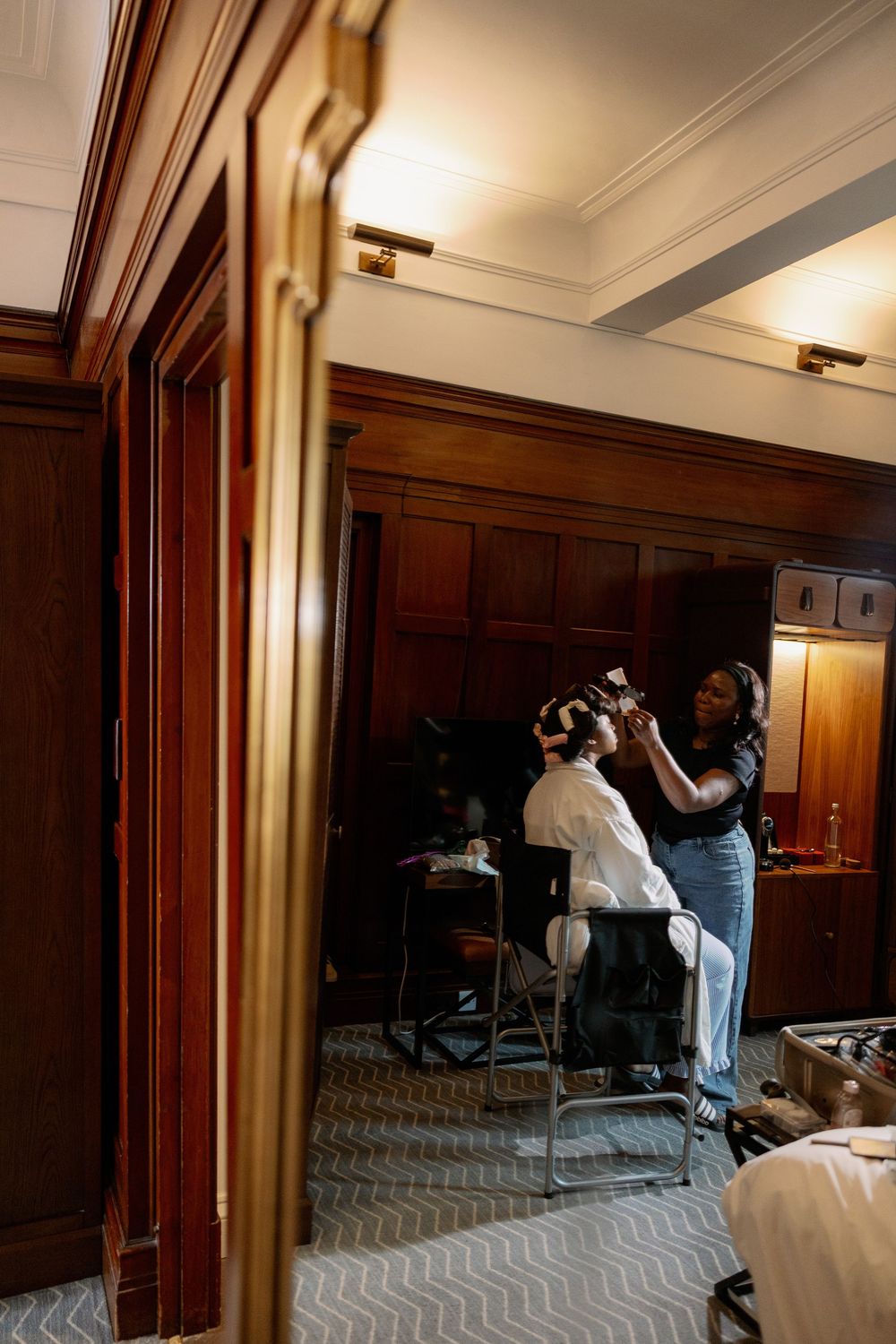 Hairstylist styling a bride's hair with rollers in an elegant wood-paneled room during wedding preparations.