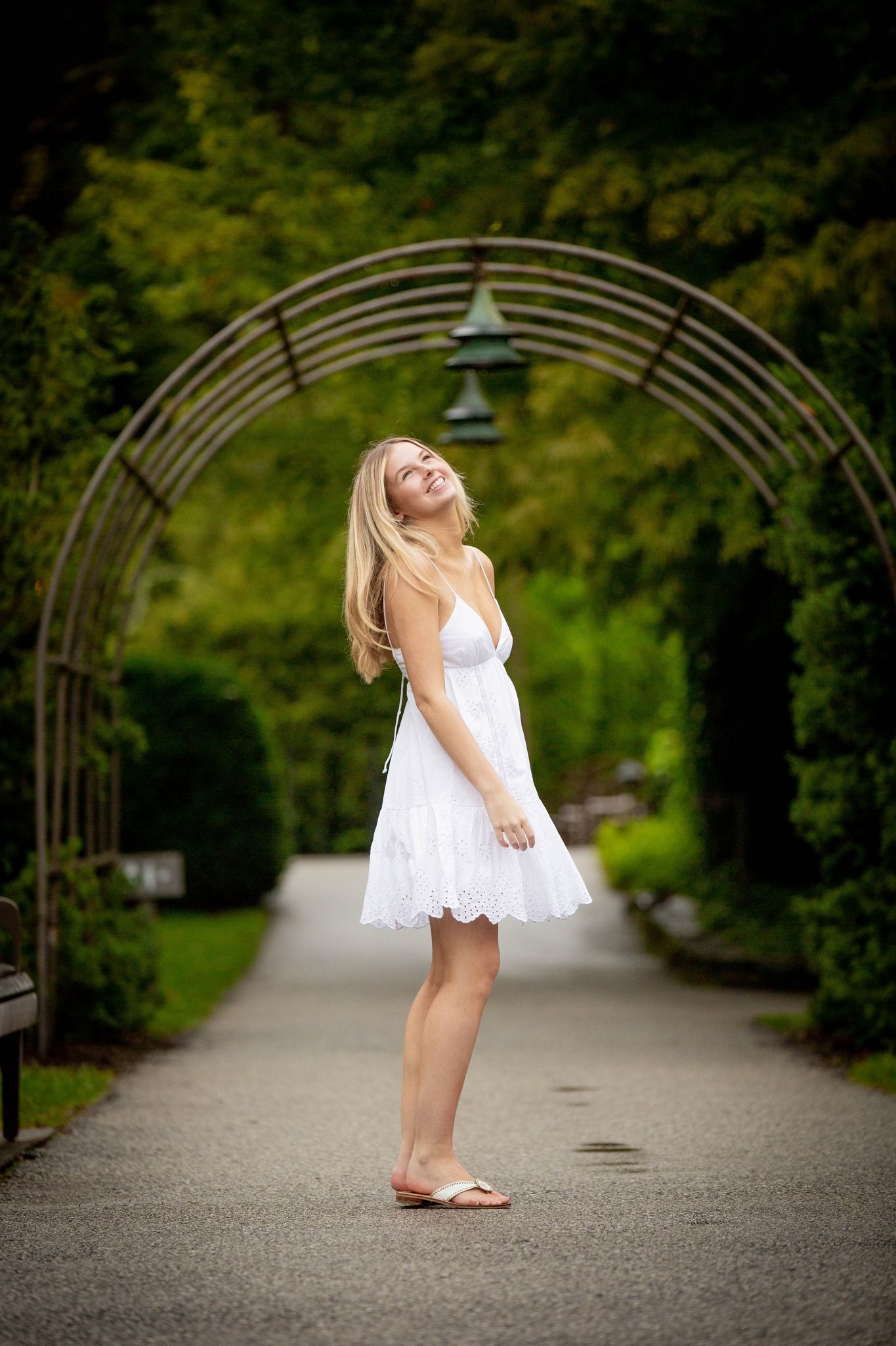 Young blonde woman in white sundress standing on garden path under decorative metal arch.