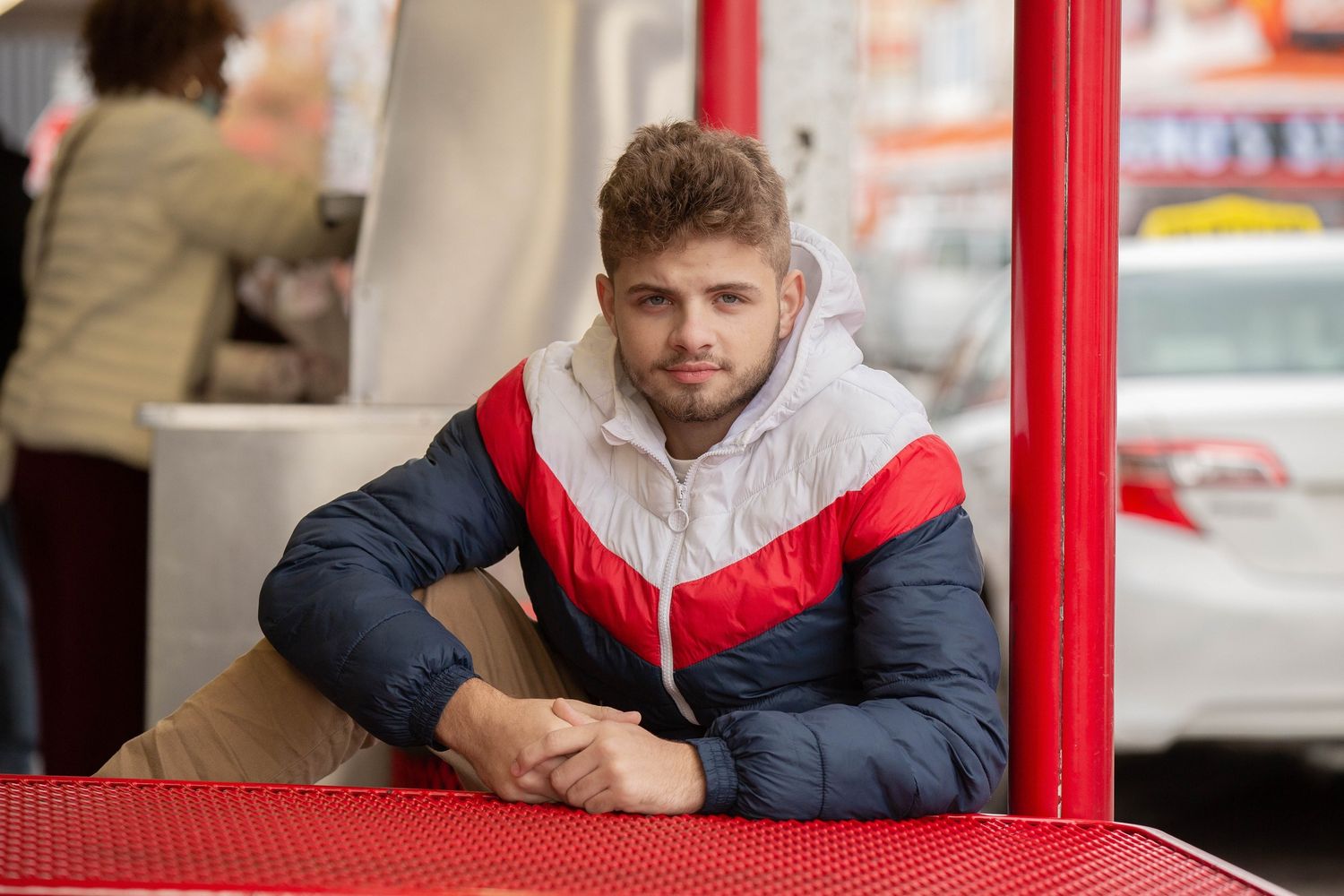 Young man in colorful winter jacket relaxes at an outdoor cafe table on a city street.