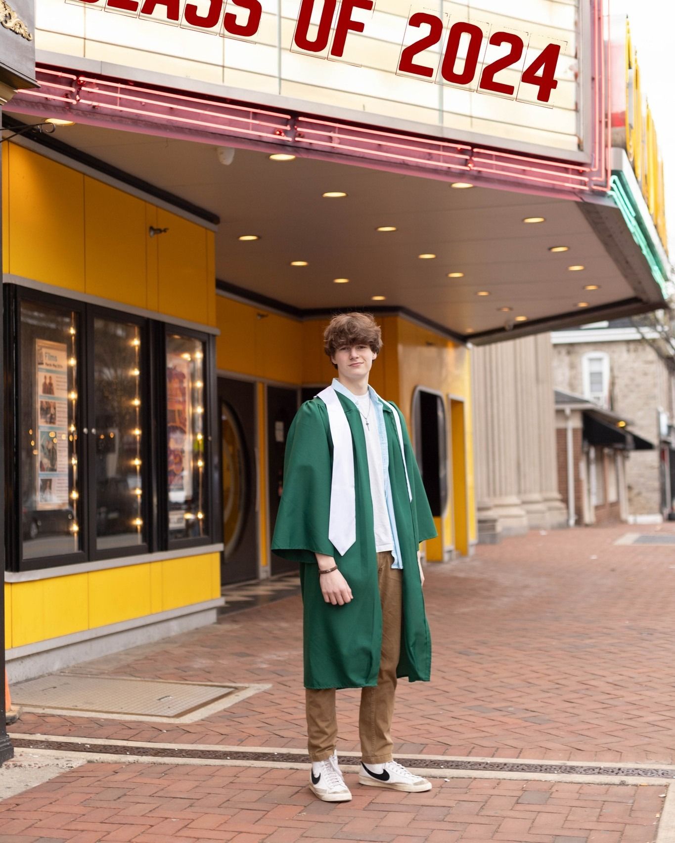 Young man in green graduation gown stands before a theater marquee reading Class of 2024.