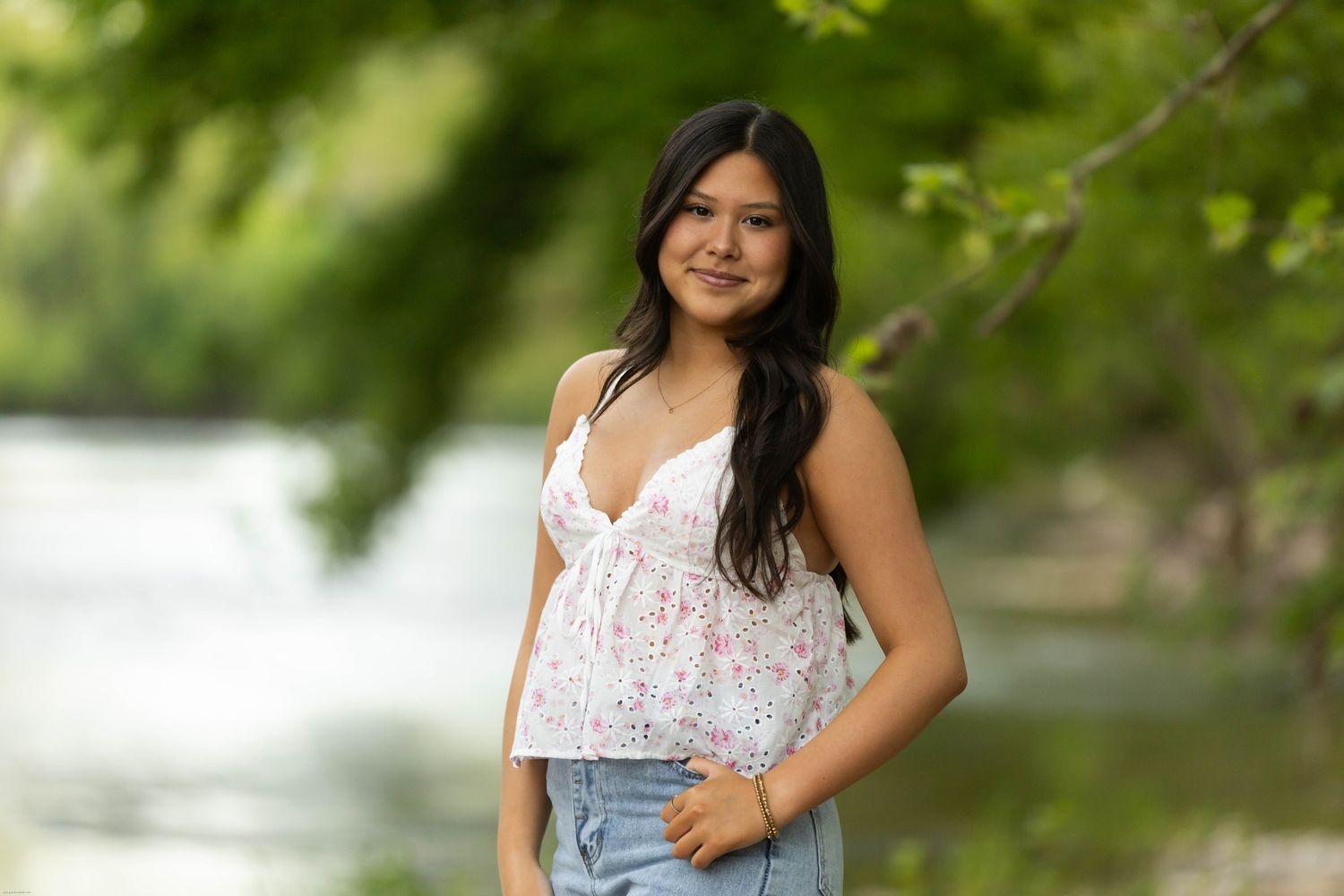 Young woman in floral top and jeans smiling outdoors near a river with lush green trees in background.
