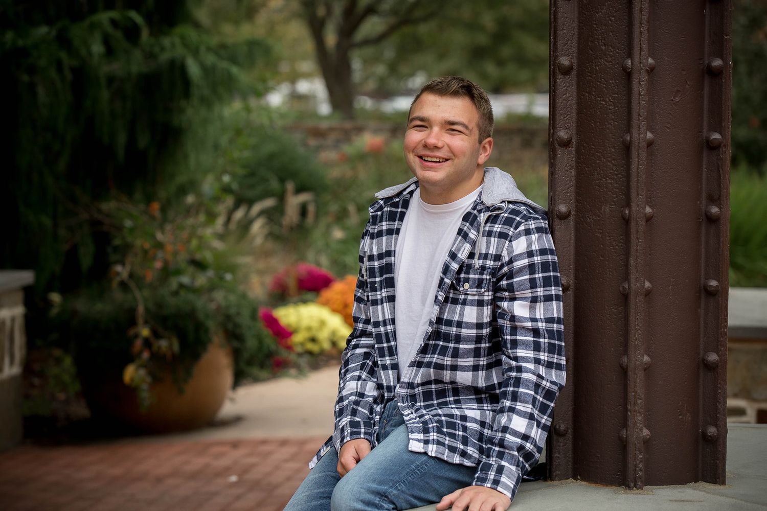 Smiling young man in plaid shirt sitting outdoors near colorful flowers and brick pillar.