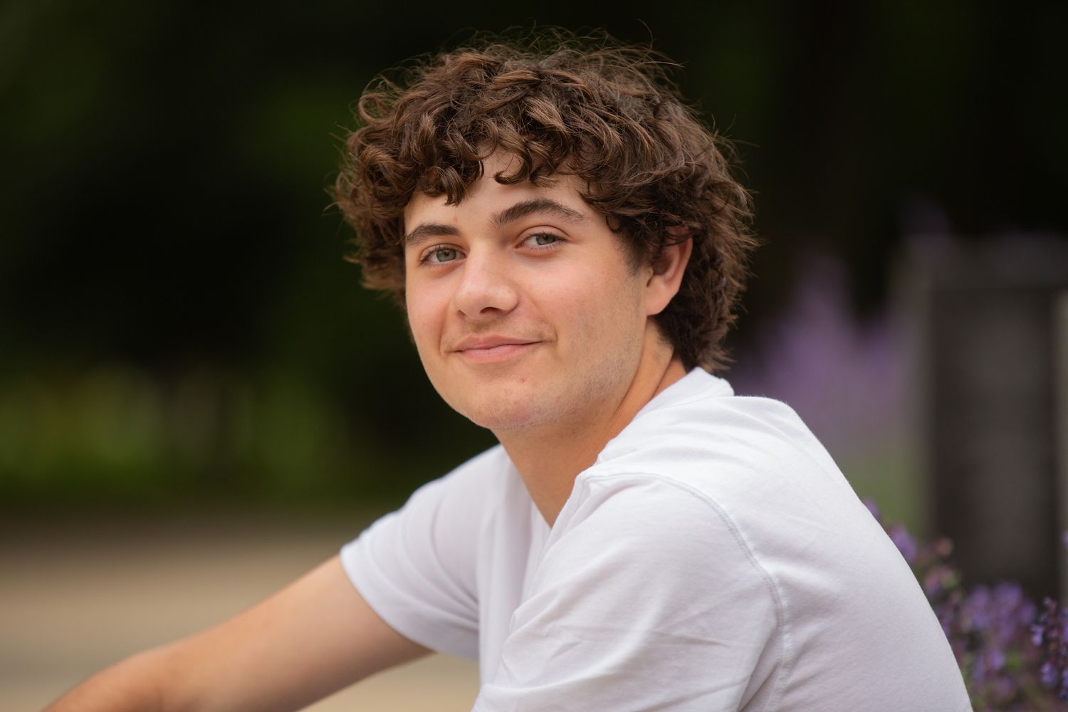 Young man with curly hair smiling outdoors in white t-shirt with green bokeh background.