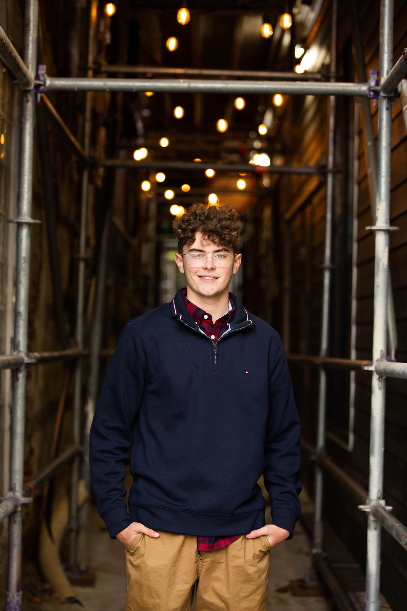Young man in navy Tommy Hilfiger pullover and khaki pants smiling in a warmly lit scaffolding corridor.