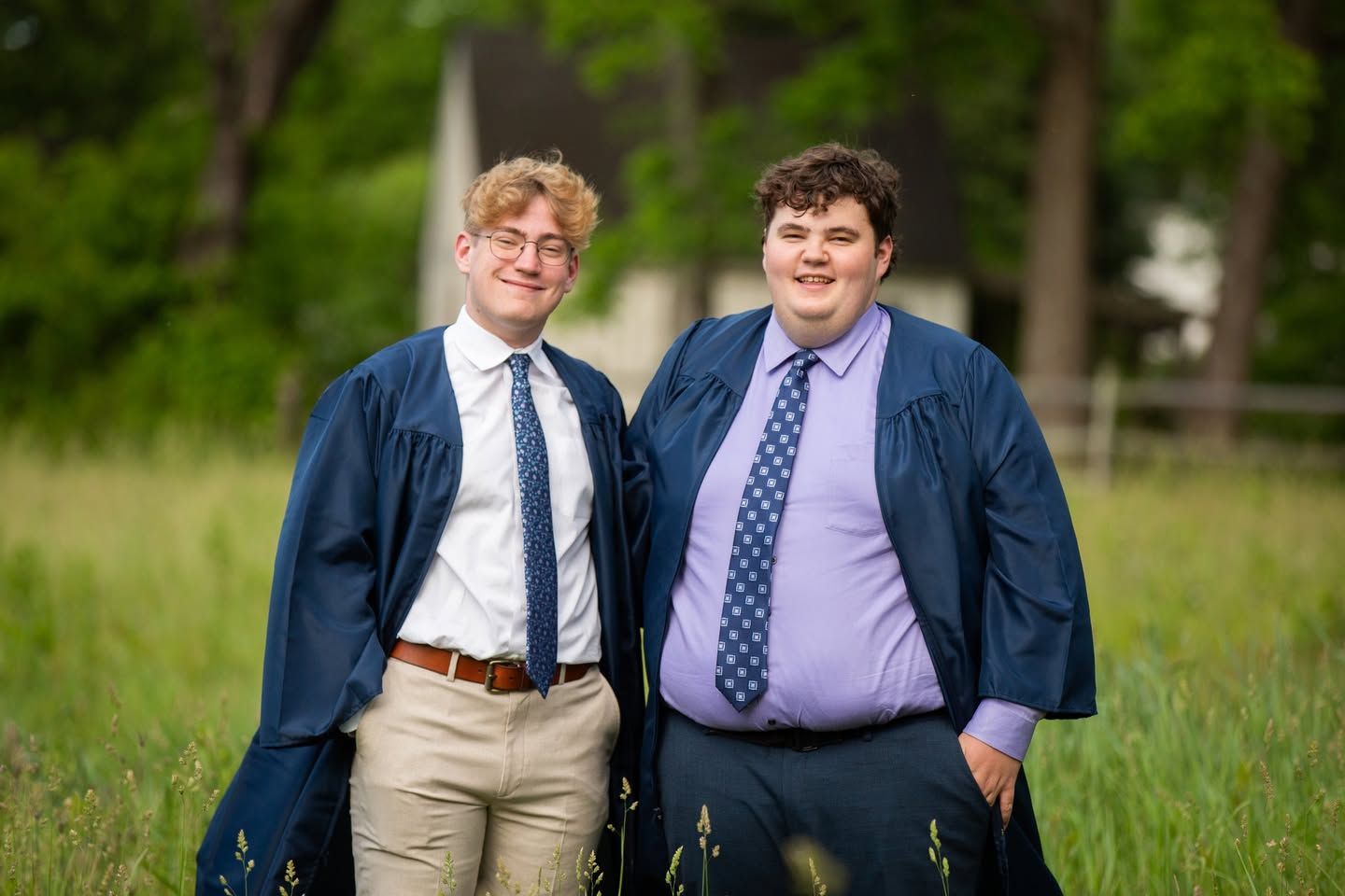 Two young men in blue graduation gowns and ties smiling outdoors in a green field.