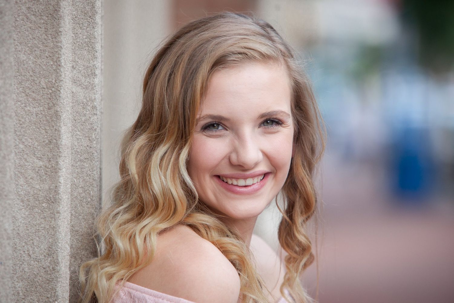 Smiling young blonde woman with wavy hair posing outdoors in a pink off-shoulder top for a portrait photo.