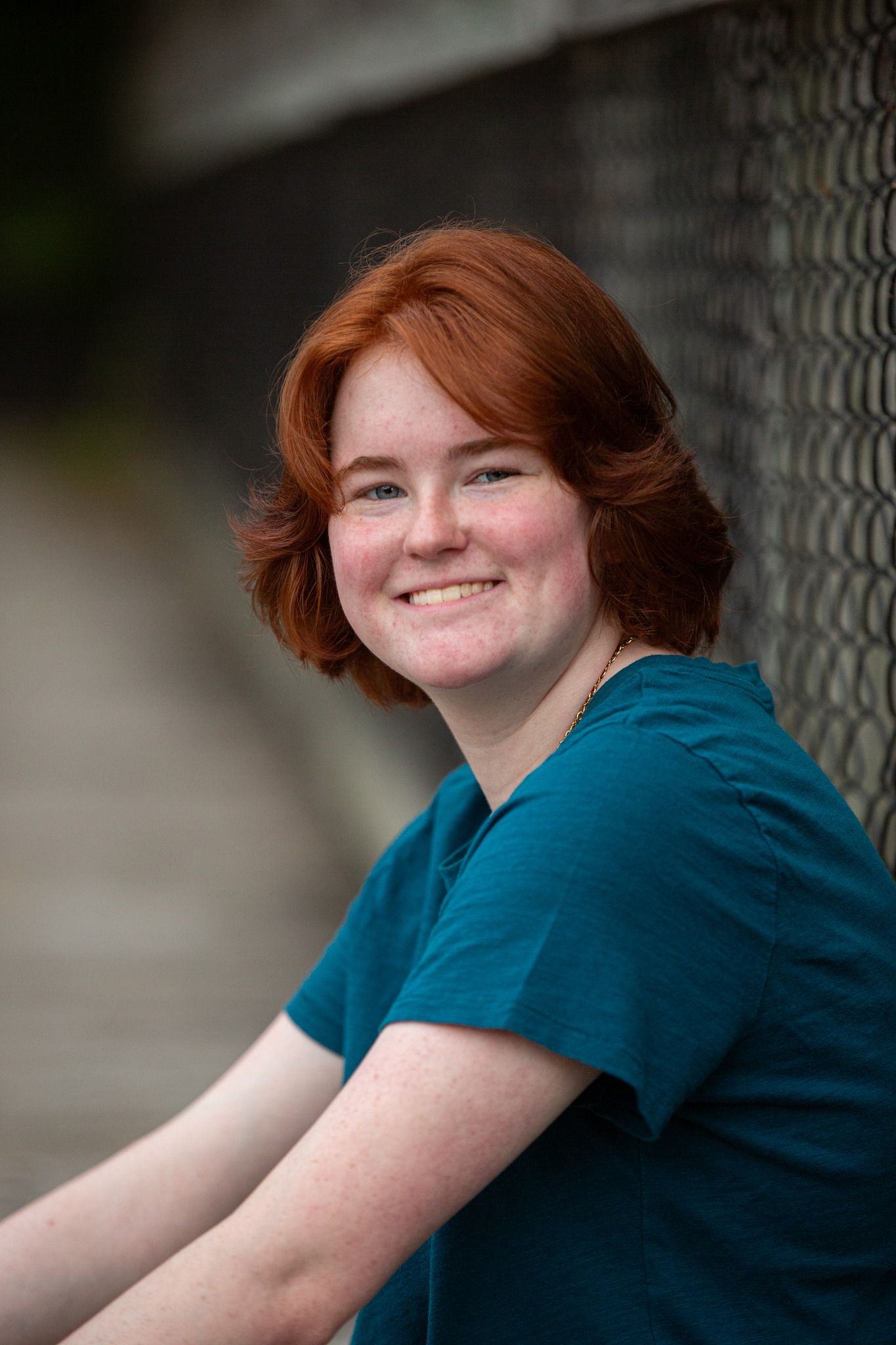 Smiling young woman with short red hair leans against a chain-link fence outdoors.