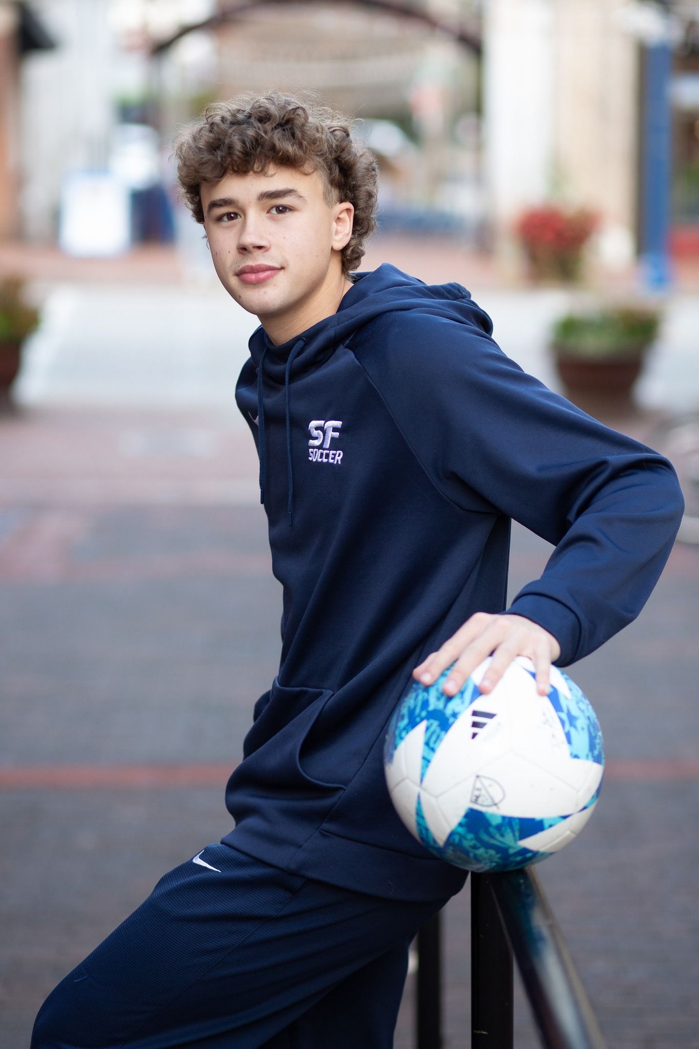 Teen soccer player in navy SF Soccer hoodie and tracksuit posing with a soccer ball outdoors.