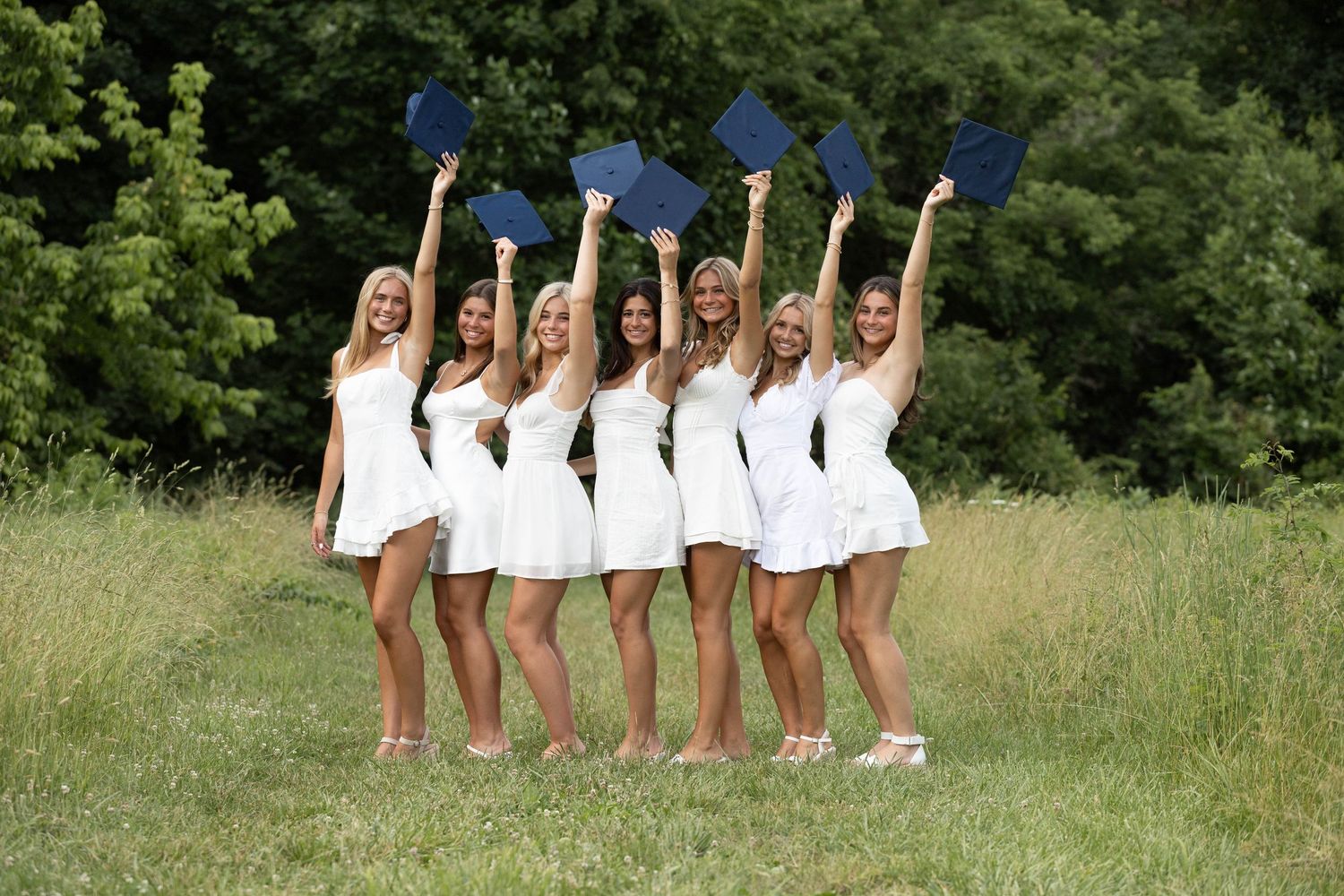 Seven young women in white dresses celebrate graduation outdoors, holding blue caps raised in the air on a sunny day.