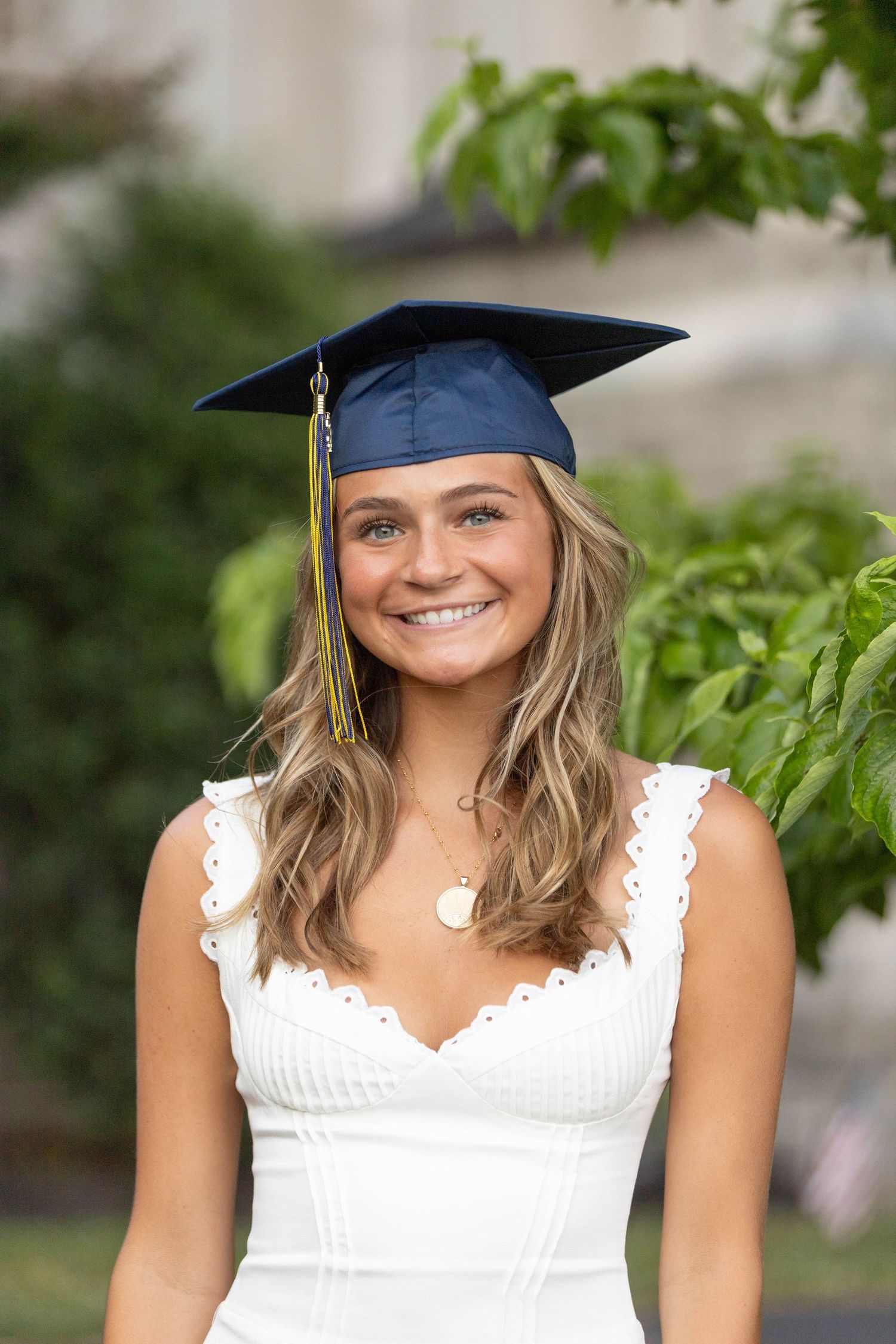 Smiling young woman wearing a navy blue graduation cap and white dress, posing outdoors for graduation photos.
