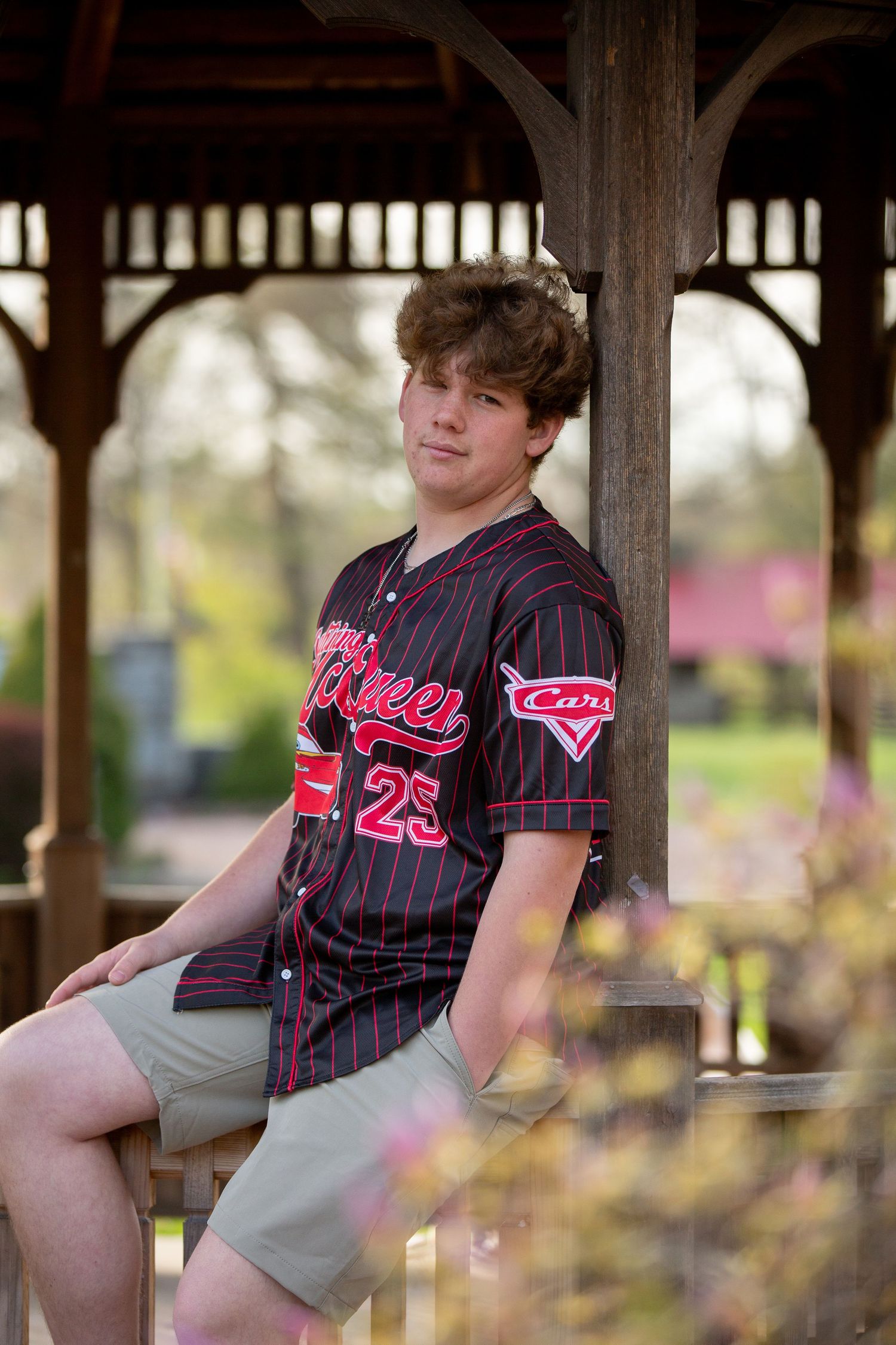 Teen boy in black pinstripe baseball jersey number 25 sitting in a wooden gazebo outdoors.