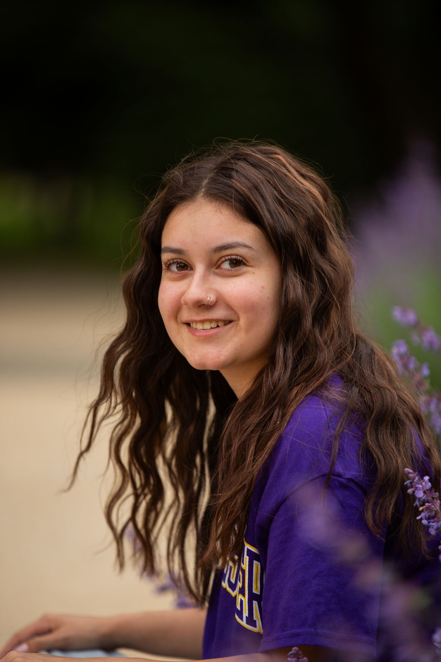 Smiling teenage girl with long wavy brown hair wearing a purple shirt, posing outdoors near purple flowers.