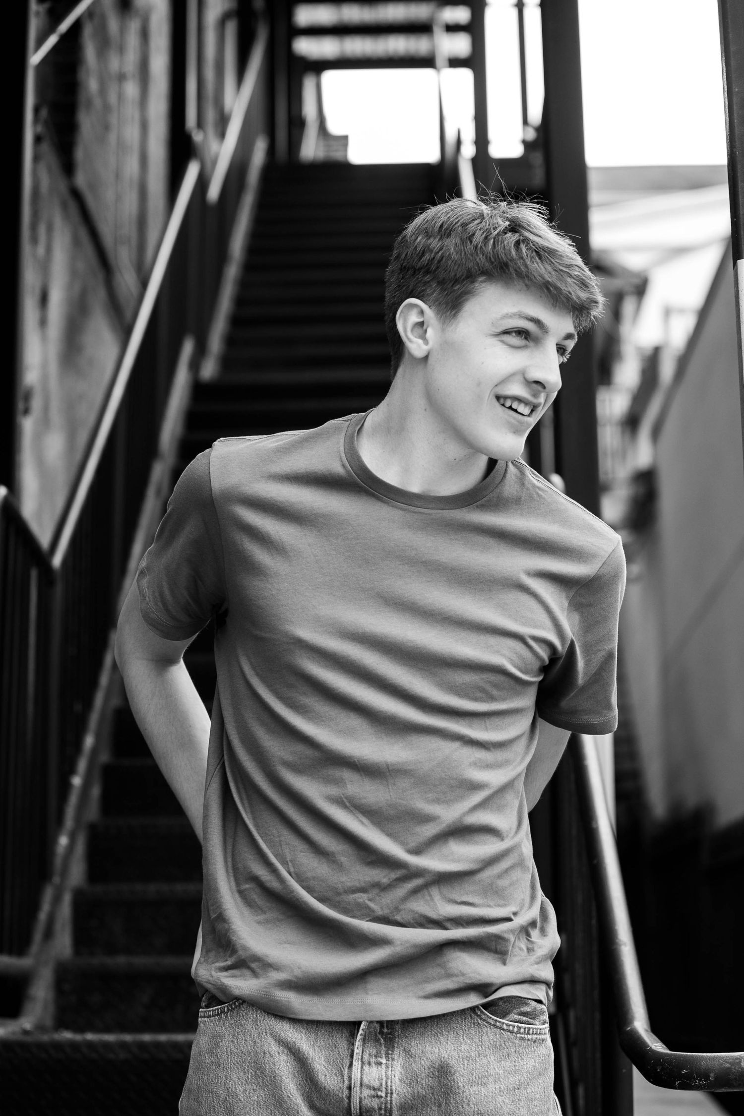 Black and white photo of a smiling young man in a t-shirt and jeans standing near metal stairs outdoors.