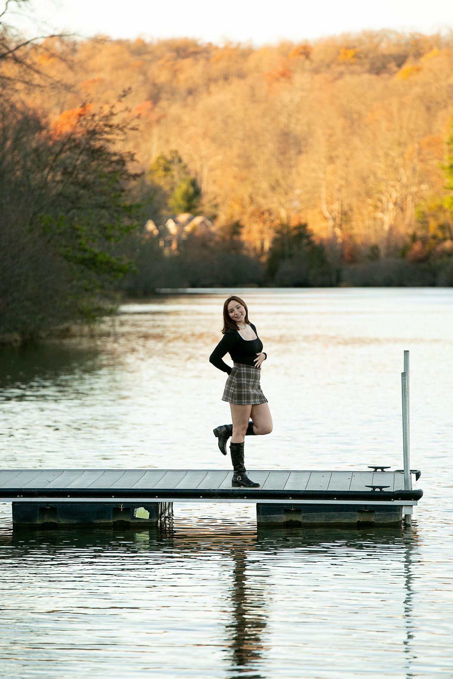Young woman in plaid skirt and black boots posing on a lake dock surrounded by autumn foliage.