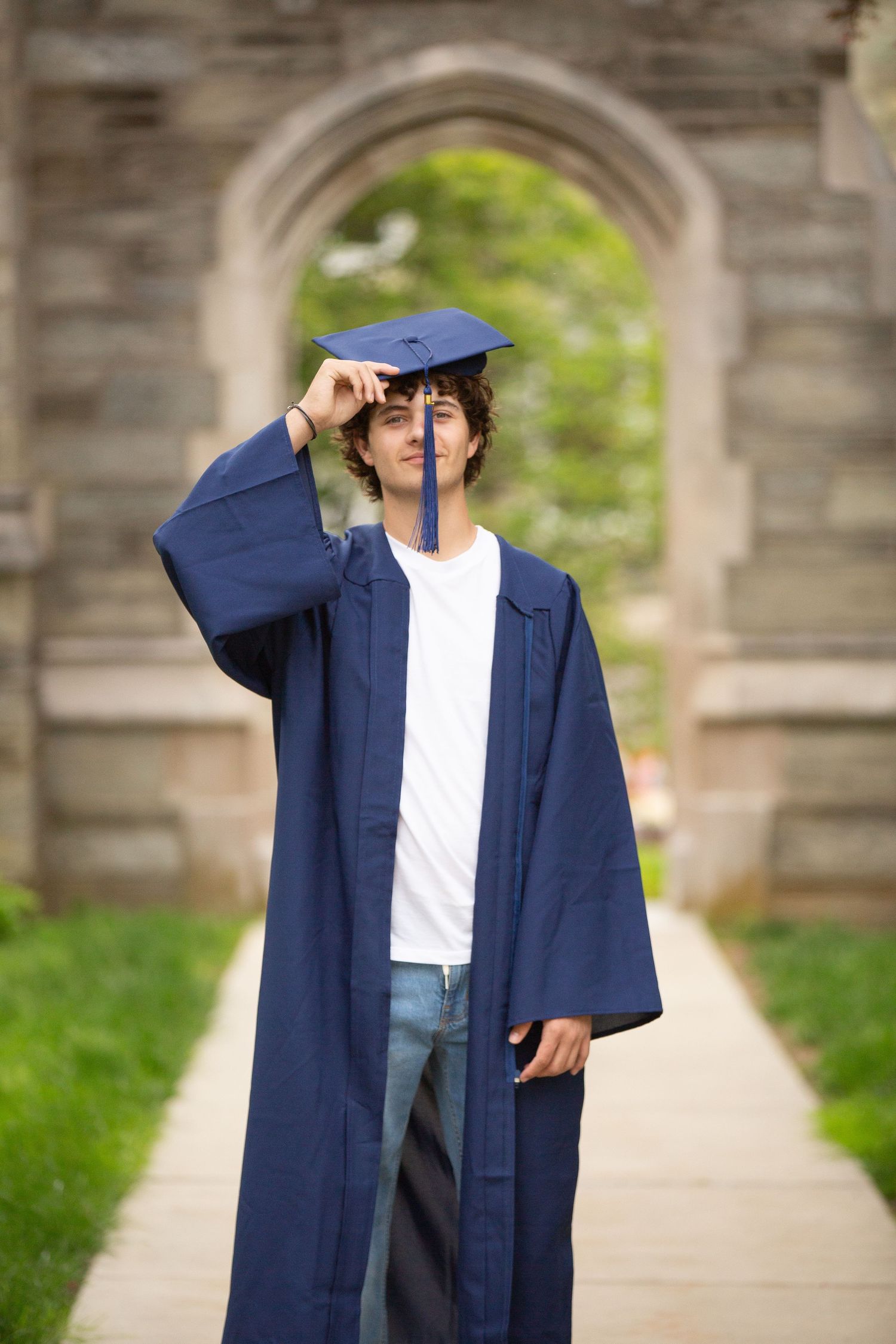Young man in navy blue graduation cap and gown posing outdoors near stone archway on campus.