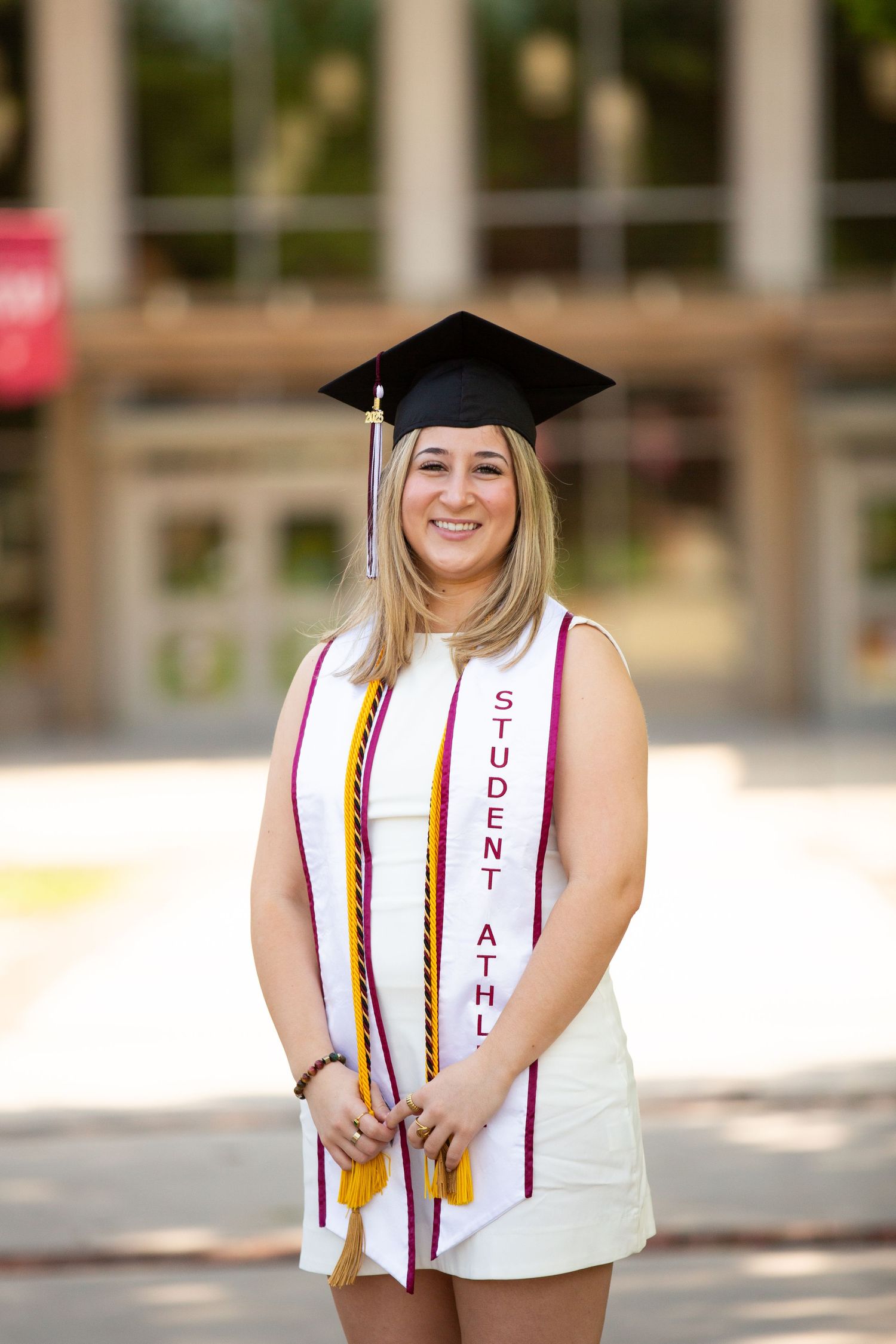 Smiling female graduate in cap, gown stole reading Student Athlete, posing outdoors on campus.