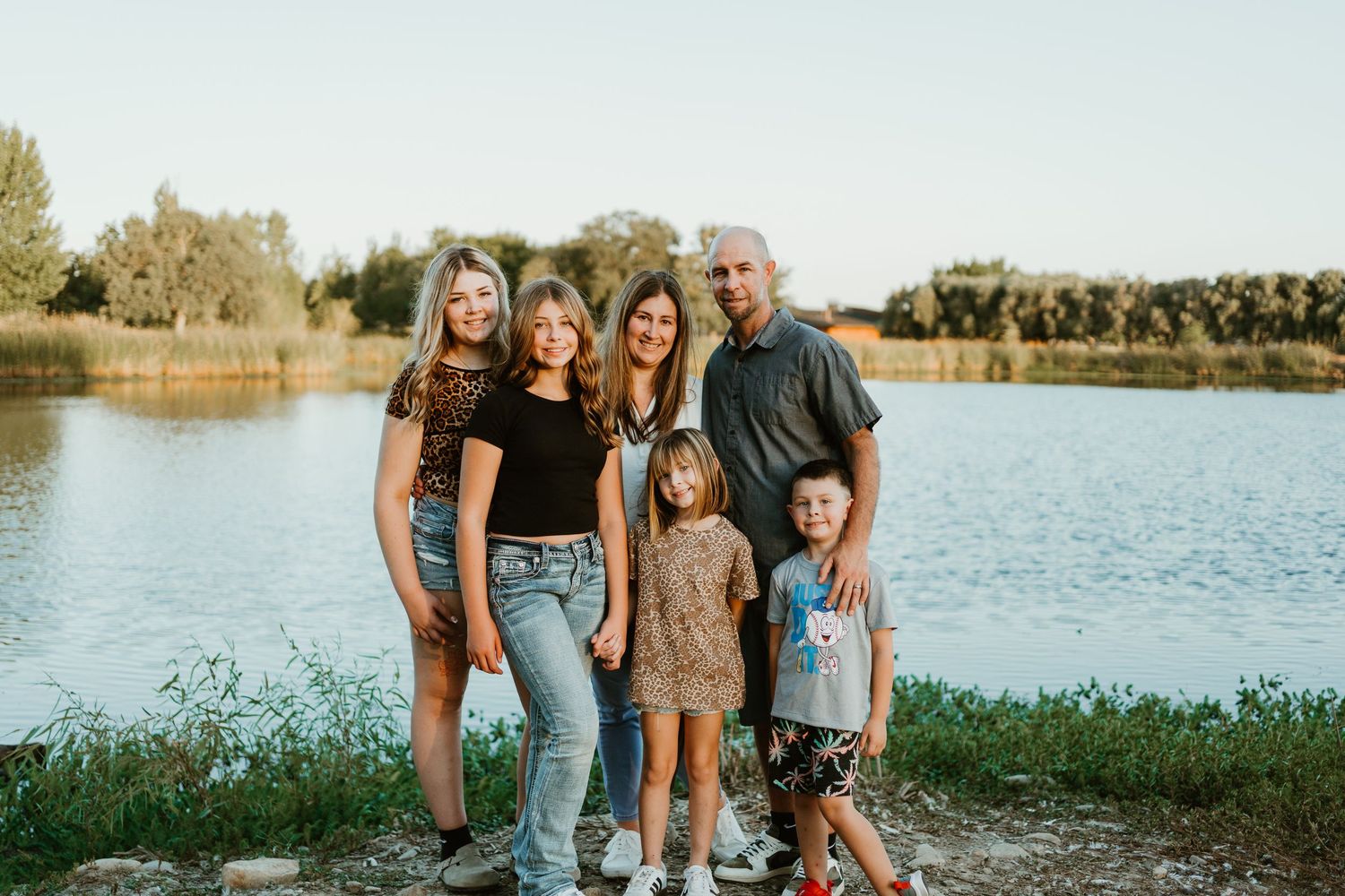 Family of six smiling together by a scenic lake surrounded by lush green trees on a sunny day.