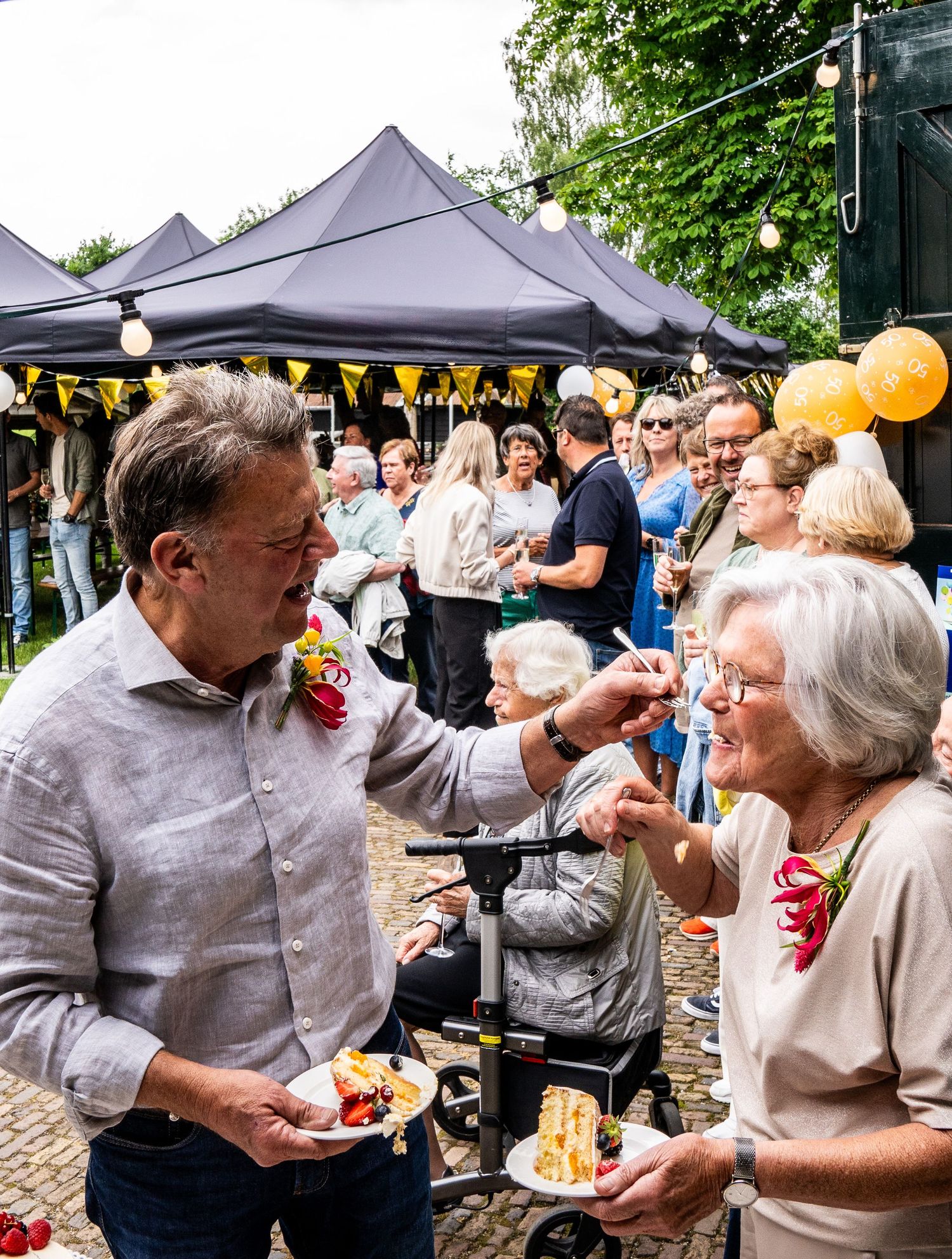 Een man deelt een taart met een oudere vrouw tijdens een buitenfeest met een menigte en zwarte tenten op de achtergrond.