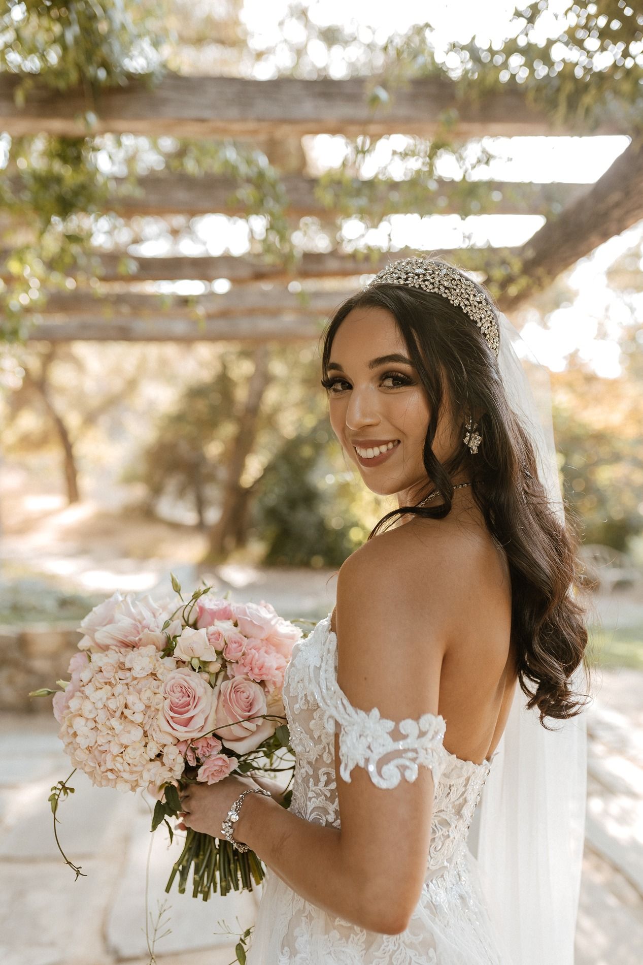 Bride in off-shoulder lace gown holding pink rose bouquet, wearing crystal tiara and veil, smiling outdoors.