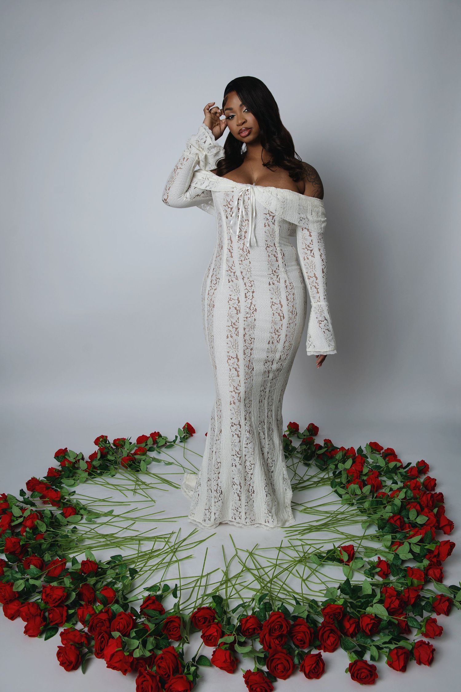 A figure in a white lace dress poses elegantly against a gray backdrop surrounded by scattered red roses on the floor.