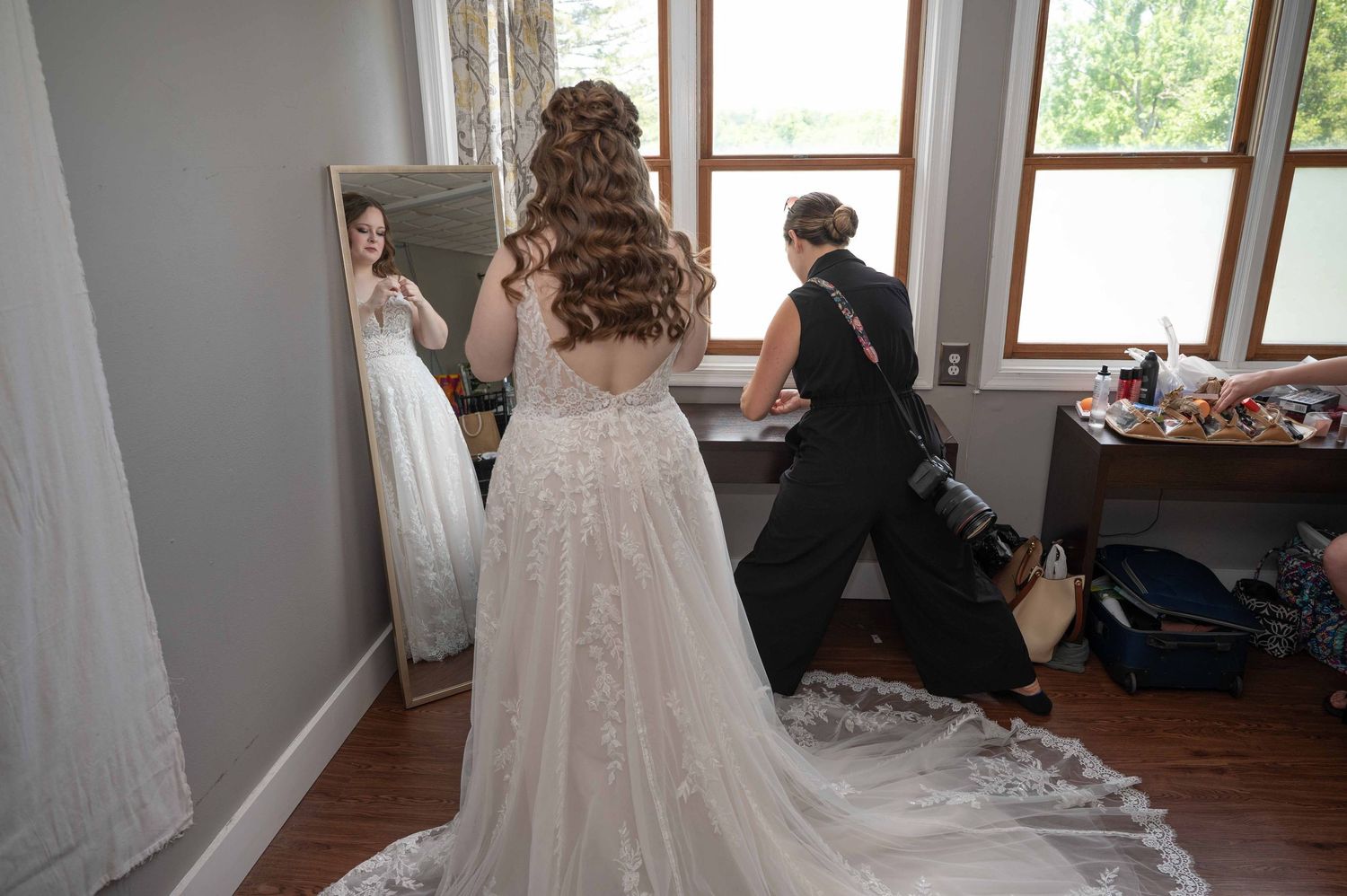 A bride in a flowing white gown gets ready in a sunlit room with large windows.
