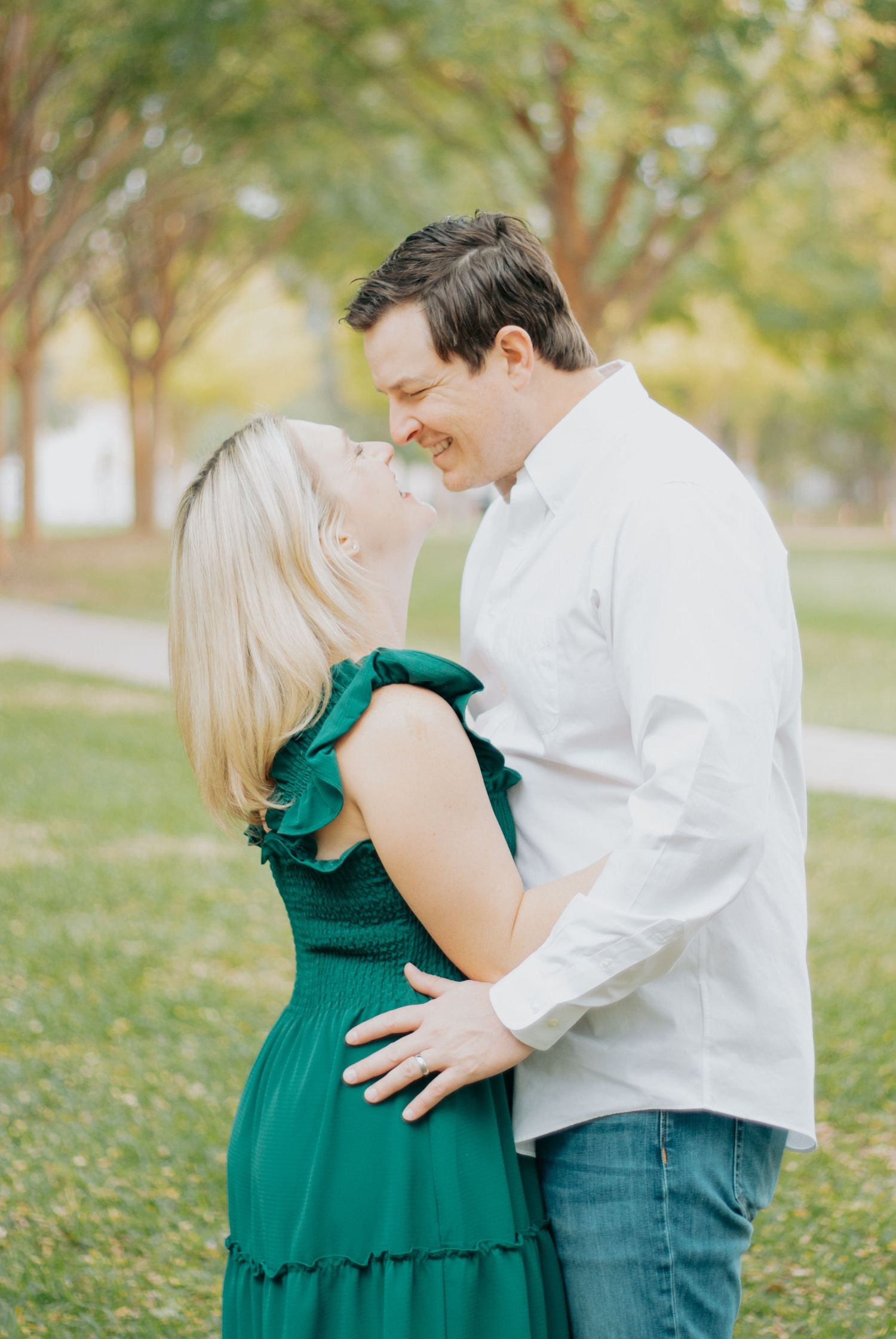A couple in a white shirt and green dress embrace lovingly in a sunlit park setting during a photo session.