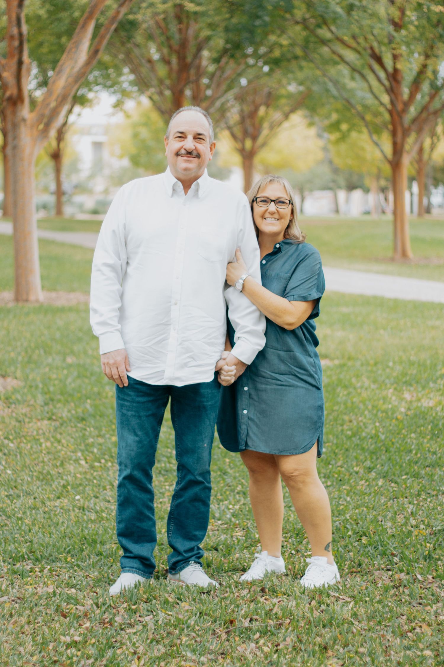 A couple stands together on green grass in a park setting wearing coordinating teal and white outfits.