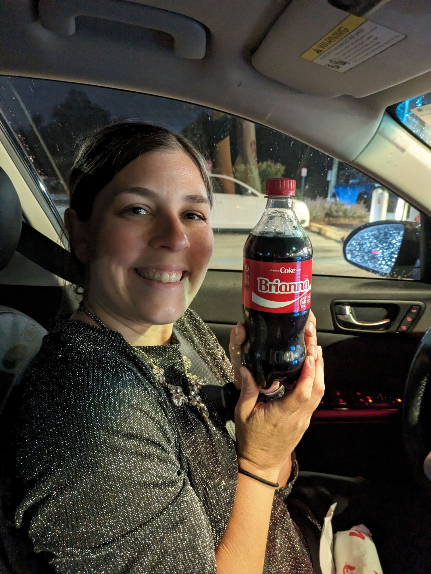 A person in a car takes selfies while holding and drinking from a glass bottle of Coca-Cola.