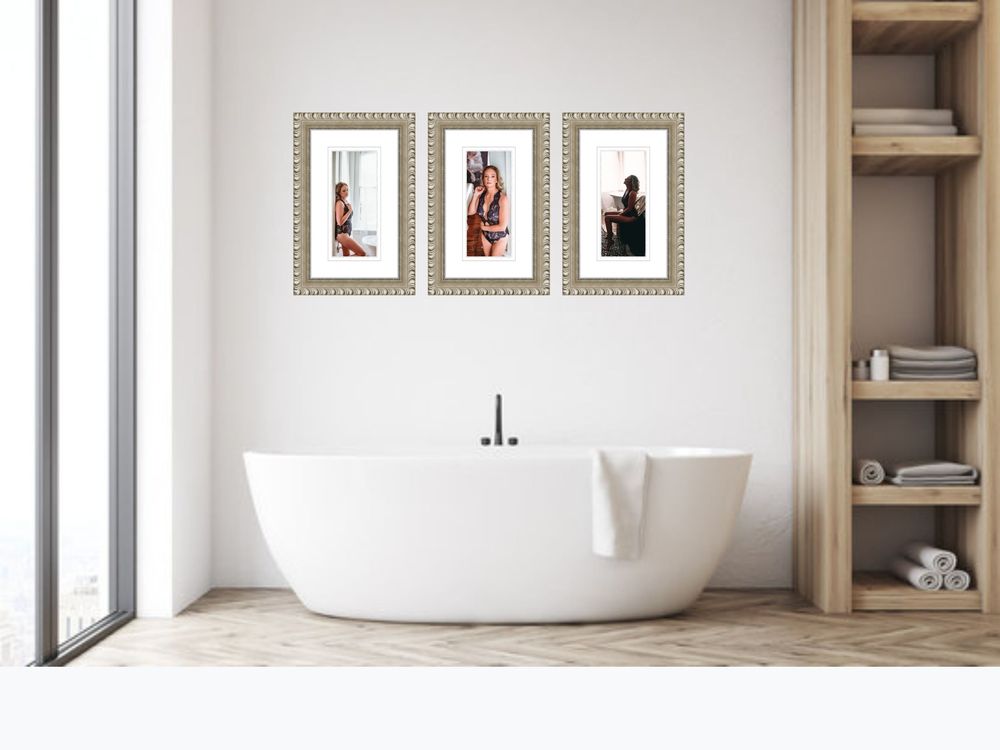 Elegant bathroom with freestanding white soaking tub, wood shelving, and three framed boudoir art prints on white wall.