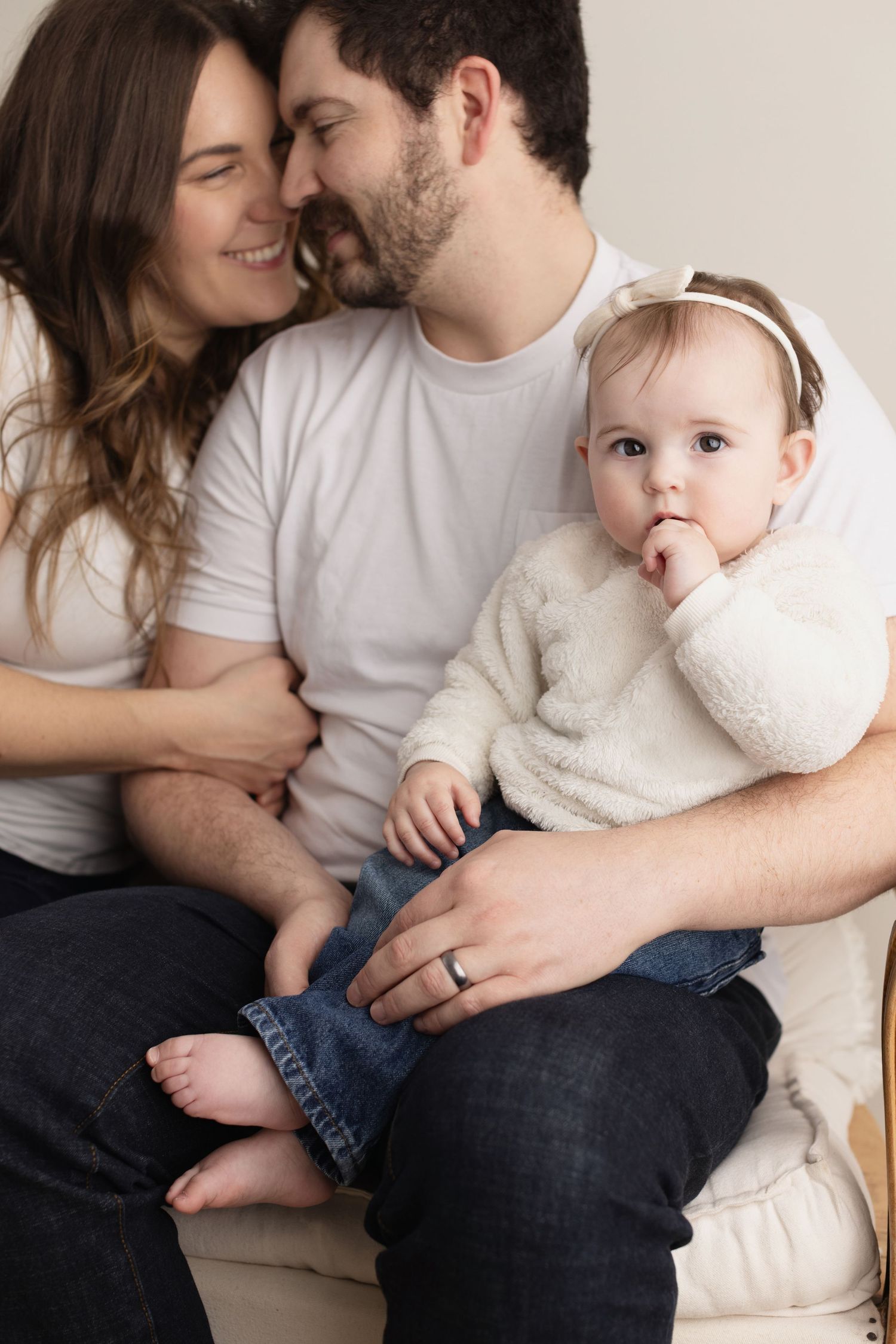 Intimate family portrait with baby in white sweater sitting together on light colored couch. Elizabeth Lang Photography Surrey