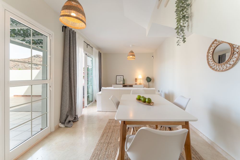 A bright, modern living room with white walls, wooden furniture, and hanging plants near large windows.