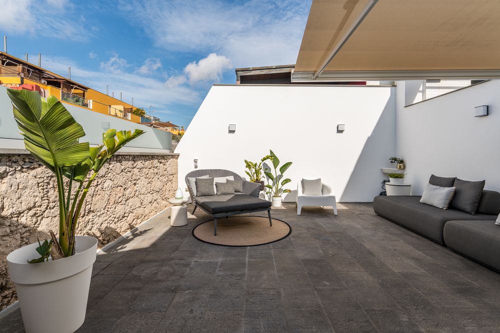 Modern rooftop terrace with grey tiles, white walls, tropical plants and comfortable outdoor furniture under blue sky.