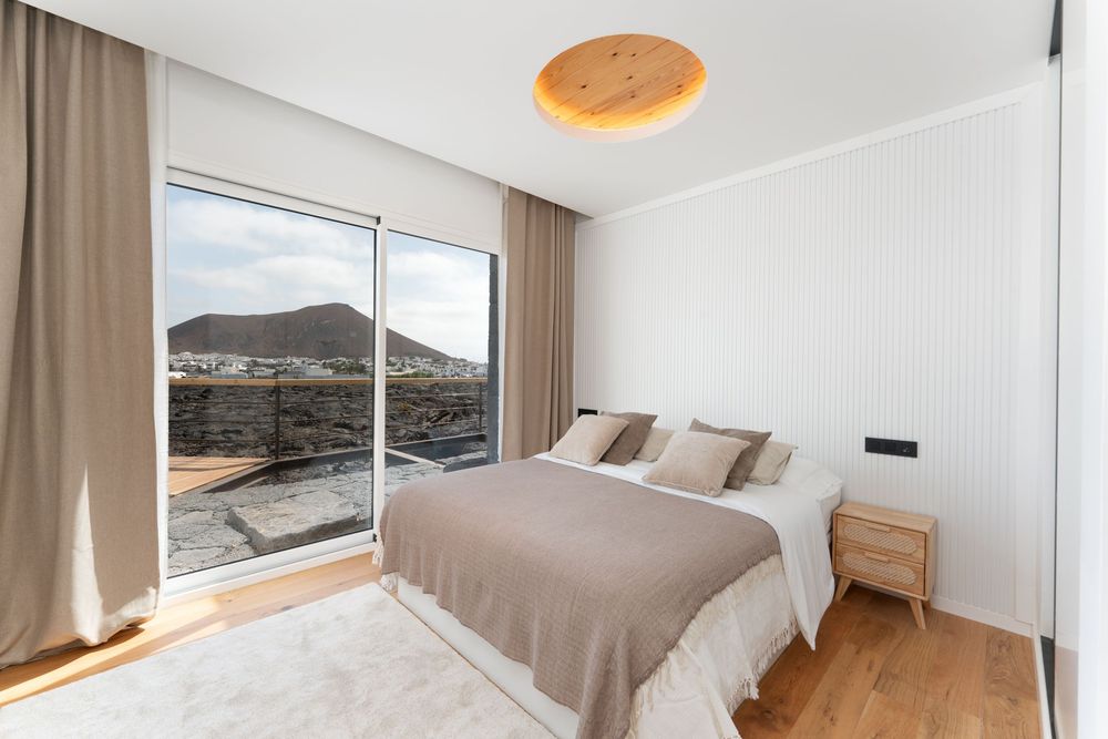 Modern bedroom with mountain views features neutral decor, wooden furniture, and floor-to-ceiling windows.