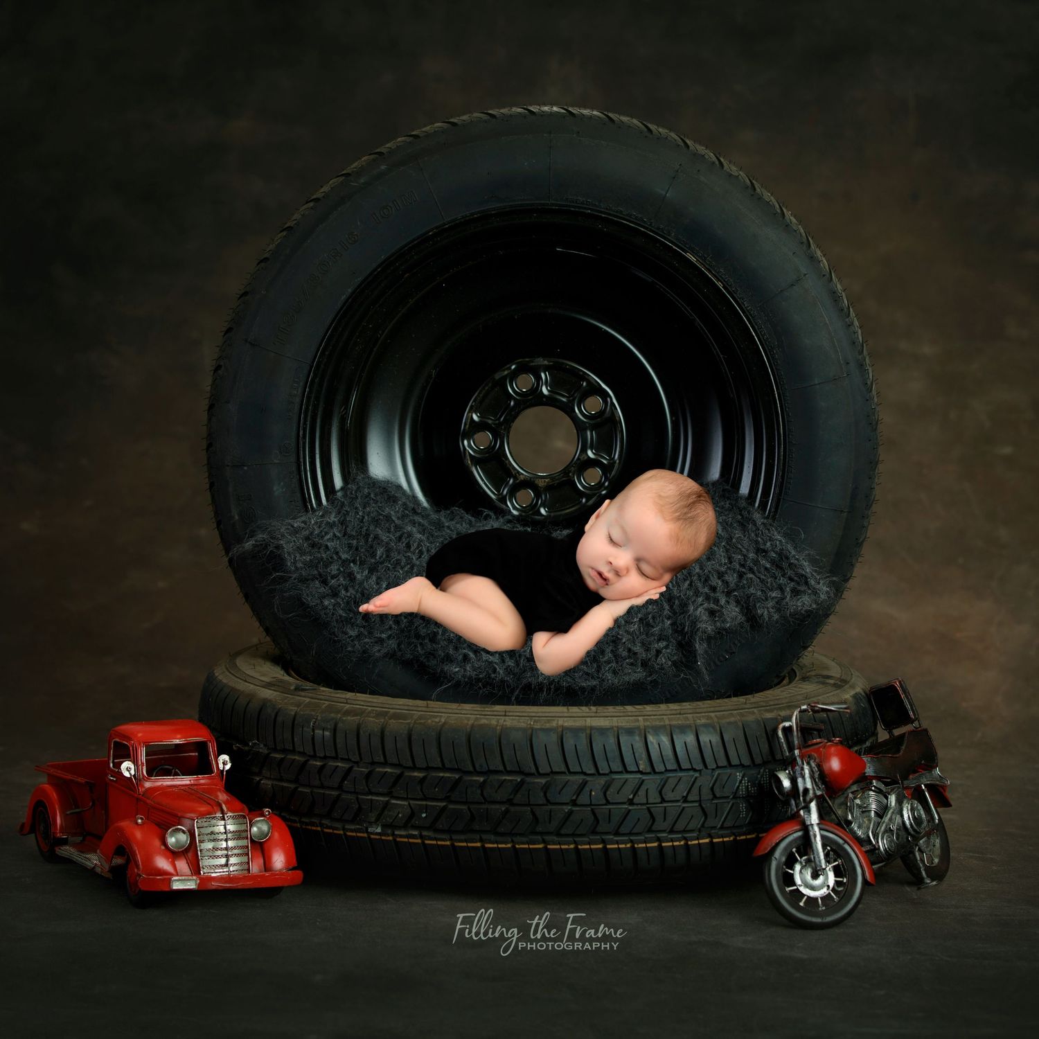 Artistic newborn photography with vintage cars and large tire props in dark studio setting.