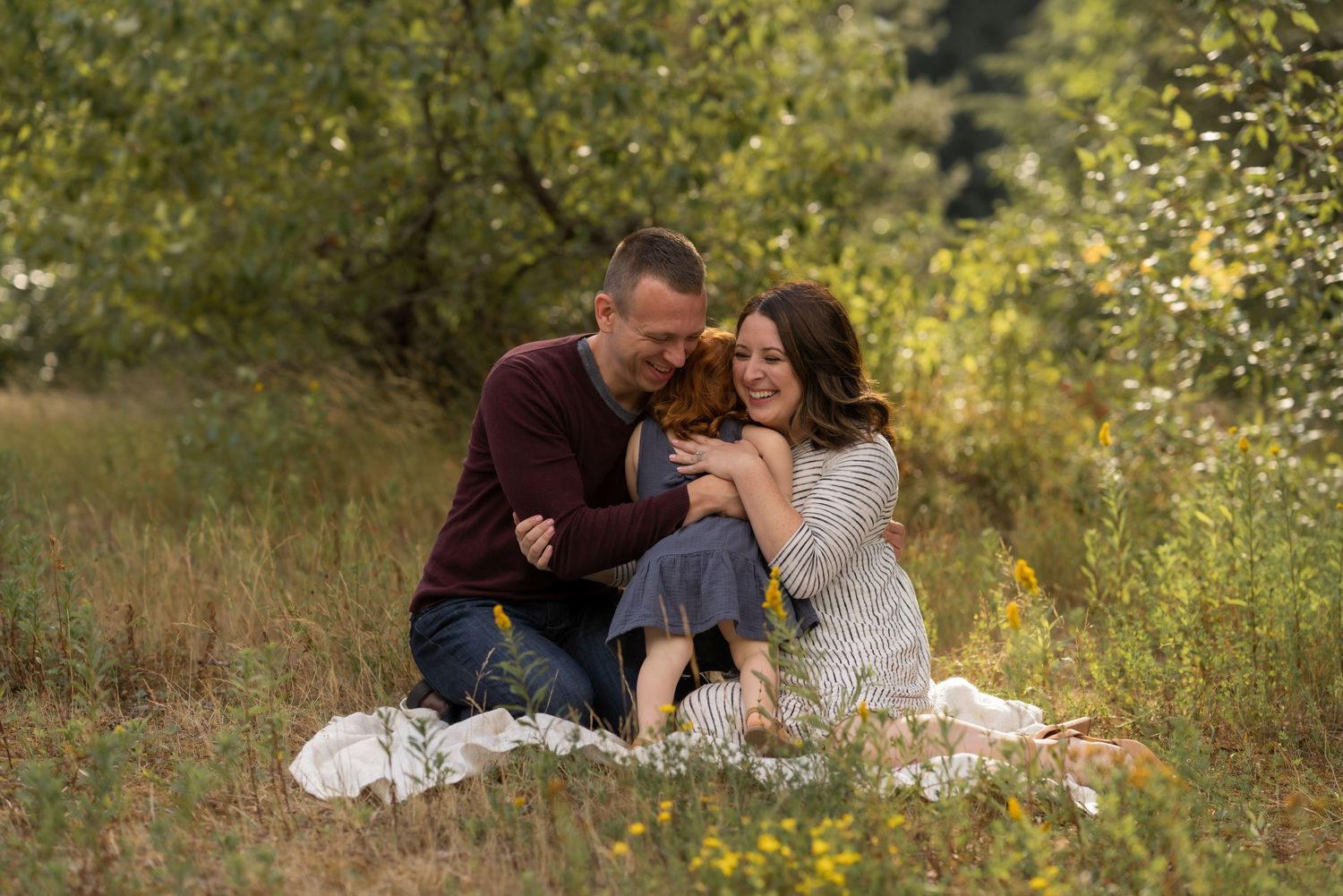 Family sitting on blanket in meadow, parents smiling and cuddling young child outdoors among wildflowers and trees.