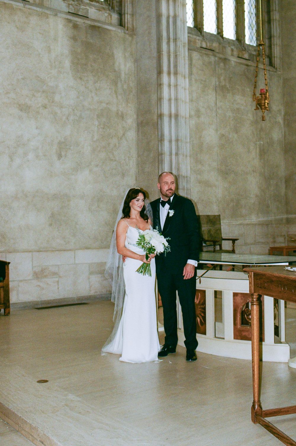 Bride and groom posing together in grand stone church after wedding ceremony, bride holding white bouquet.